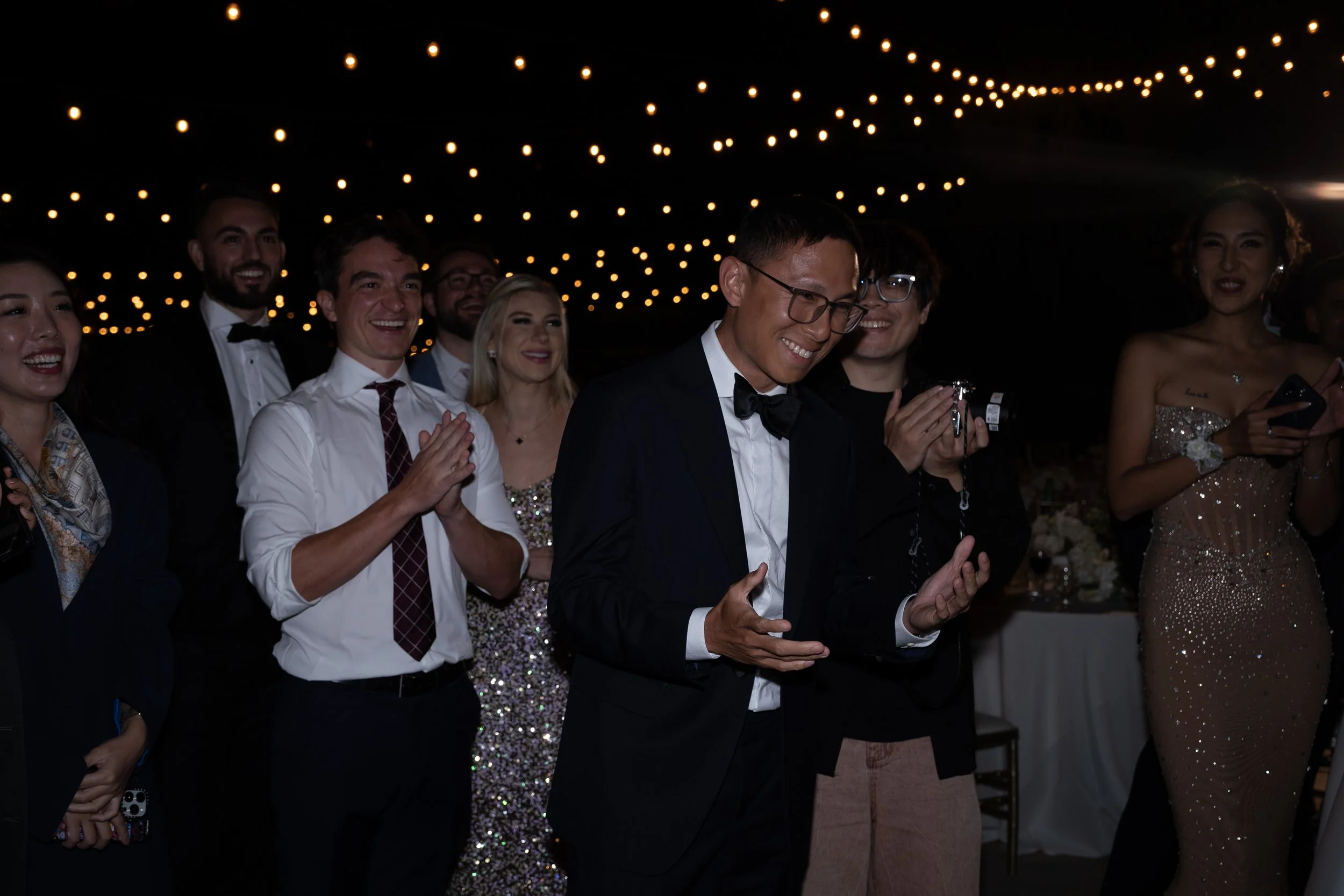 Group of people dressed in formal attire at an event with string lights overhead.