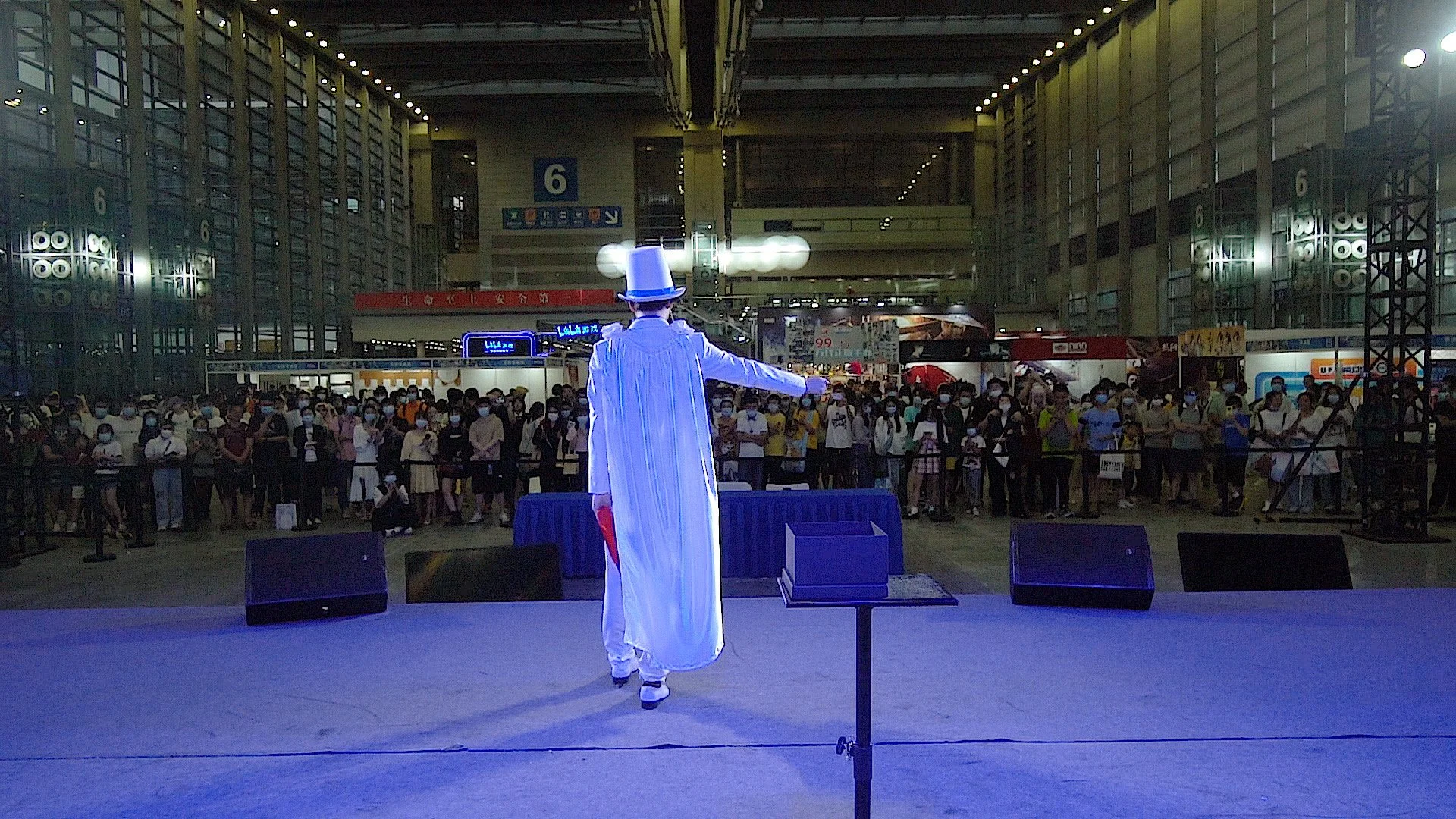 Performer wearing a white costume and blue top hat on stage, facing an audience in an indoor venue with high ceilings and glass walls.