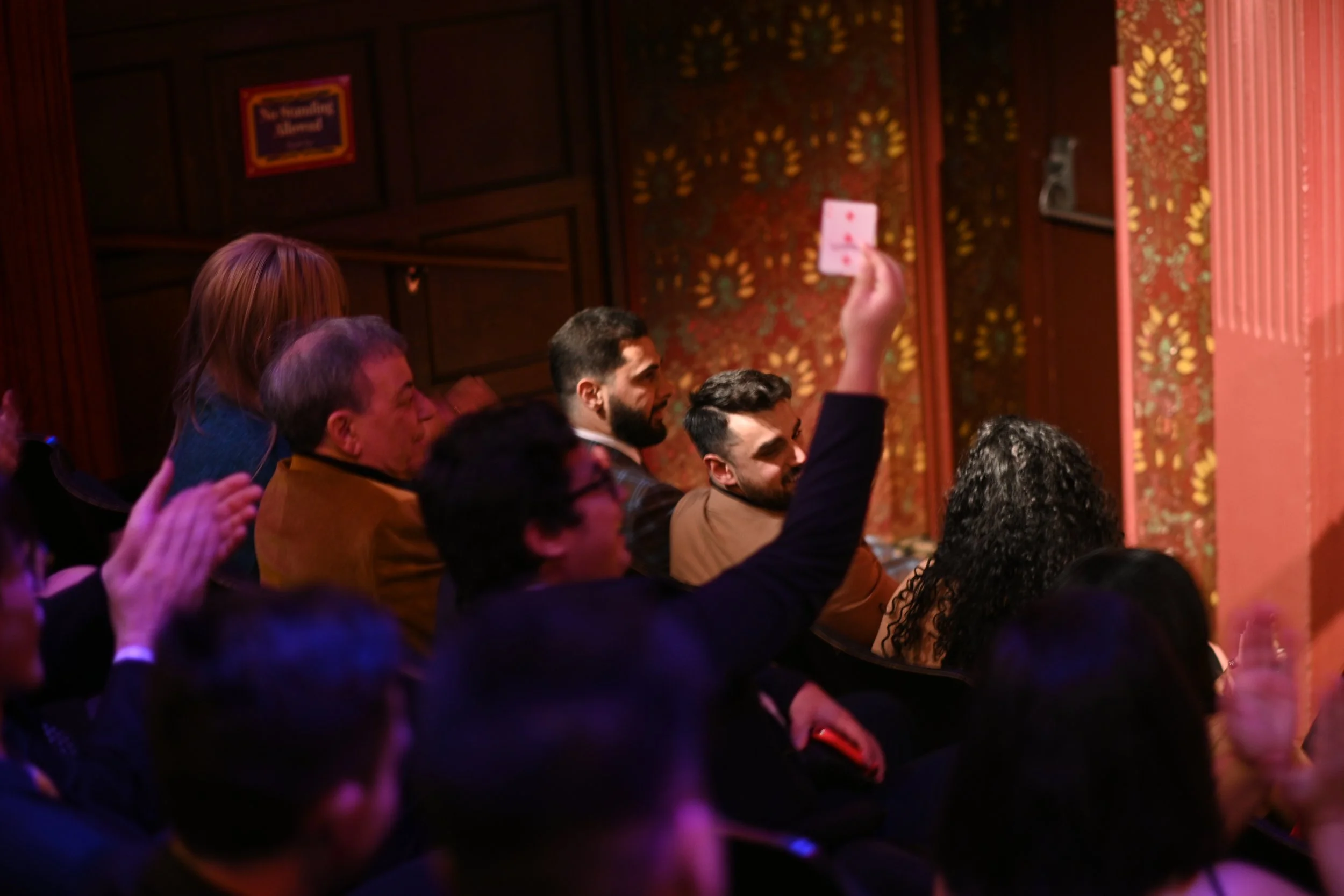 A group of people seated in a dimly lit auditorium, with some holding up phones, in front of a wall decorated with ornate wallpaper and a sign that reads "The Recording Museum."