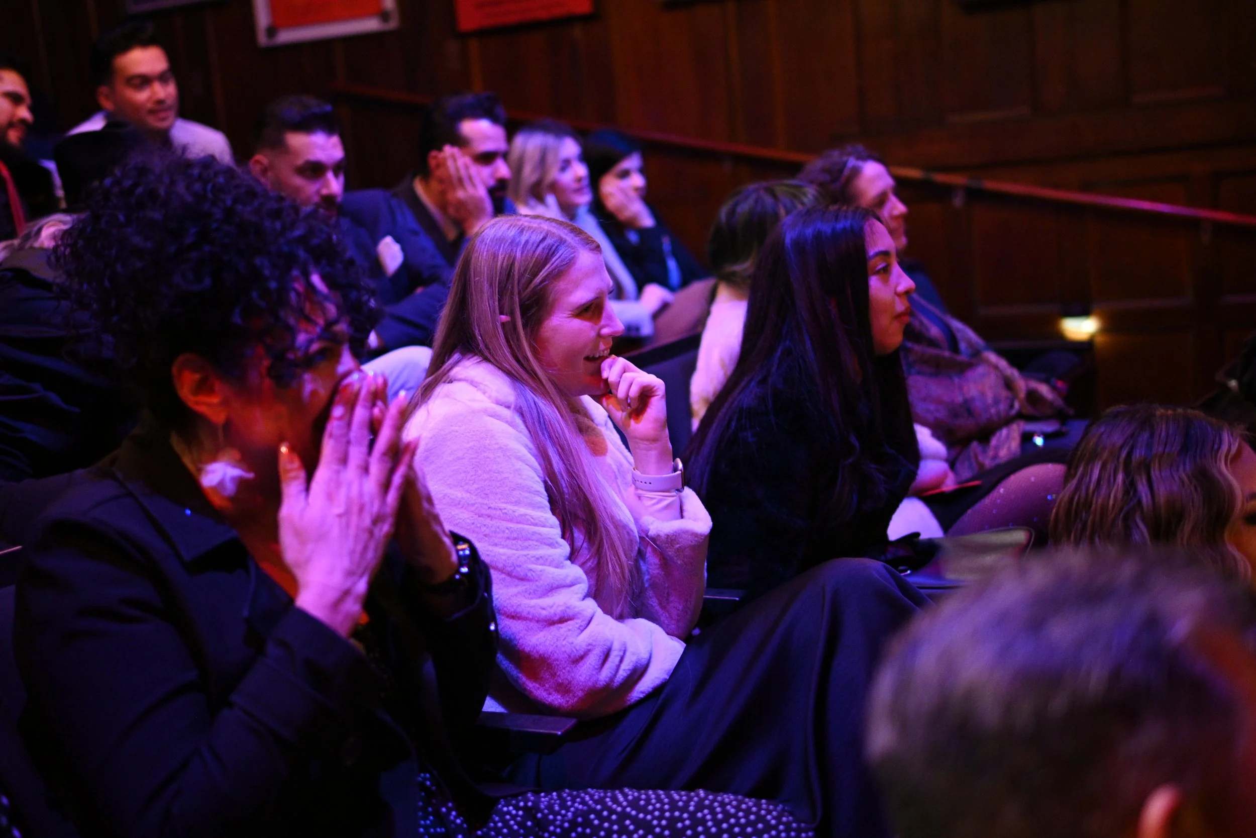 People sitting in dark room watching a performance, with a mix of ethnicities, some smiling and others focused, under dim, colorful lighting.