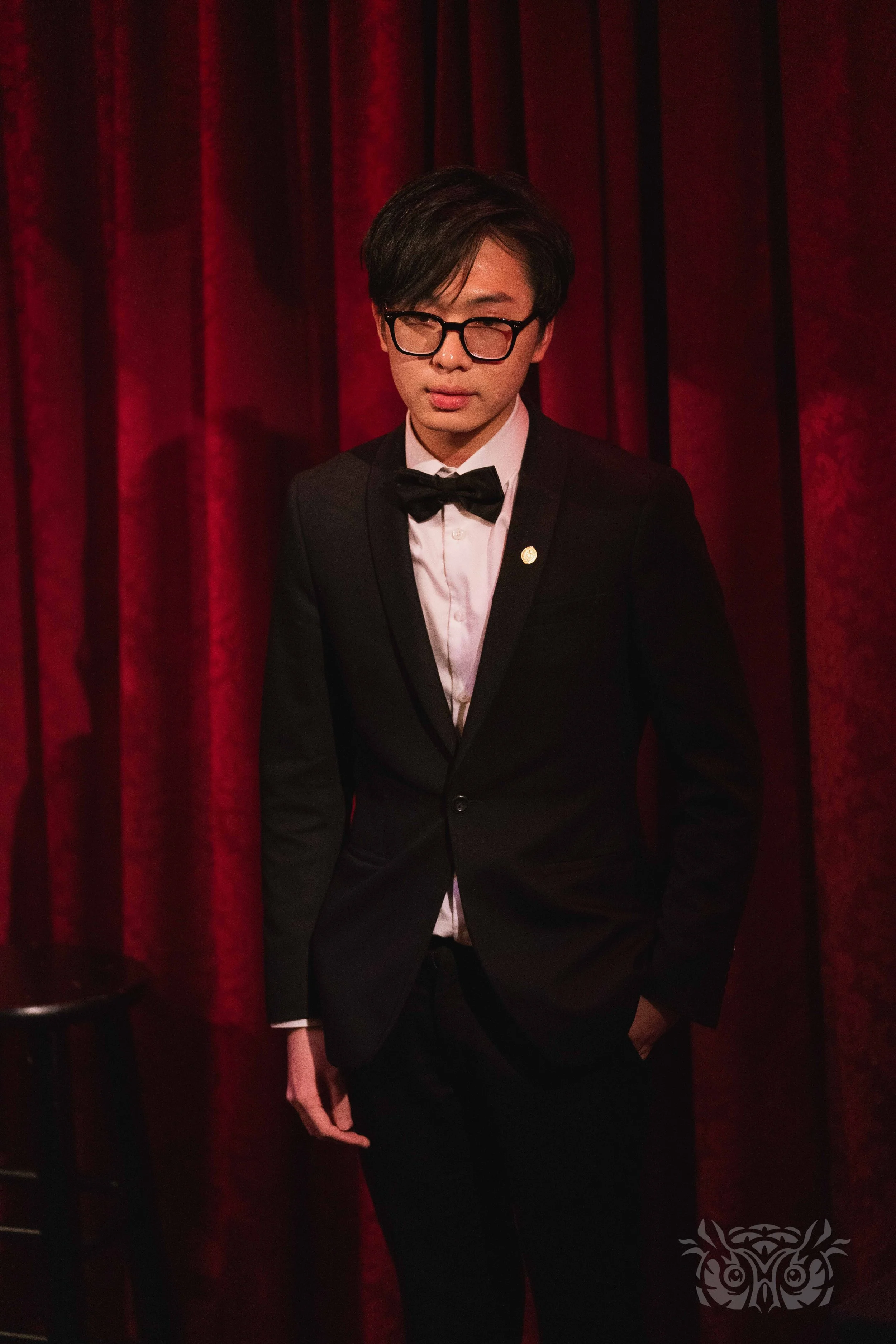 A young man in a black tuxedo, white shirt, and black bow tie standing in front of red velvet curtains.