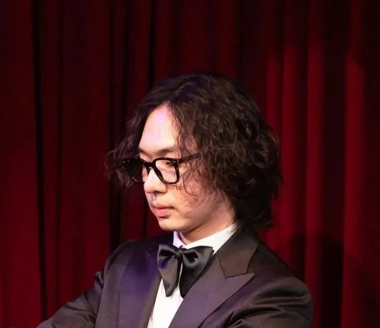 A man wearing glasses and a tuxedo with a bow tie, standing in front of red curtains.