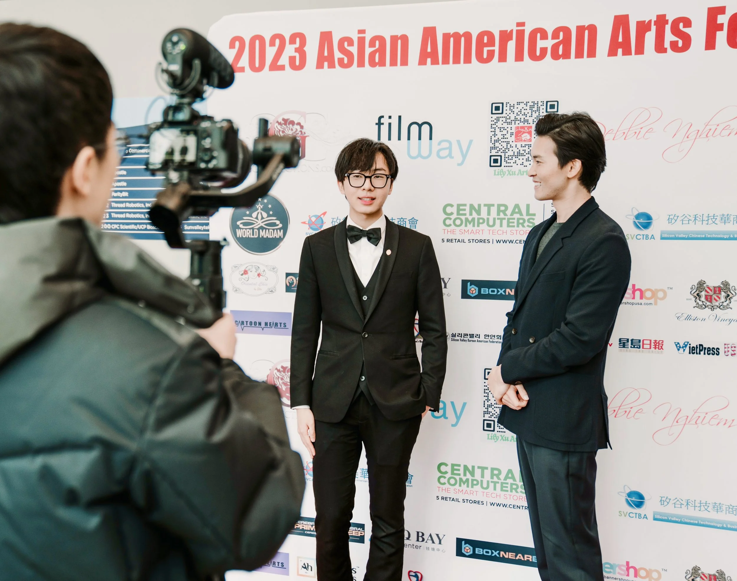 Two young men in suits and glasses being filmed at the 2023 Asian American Arts Festival, with a backdrop displaying various sponsor logos and event information.