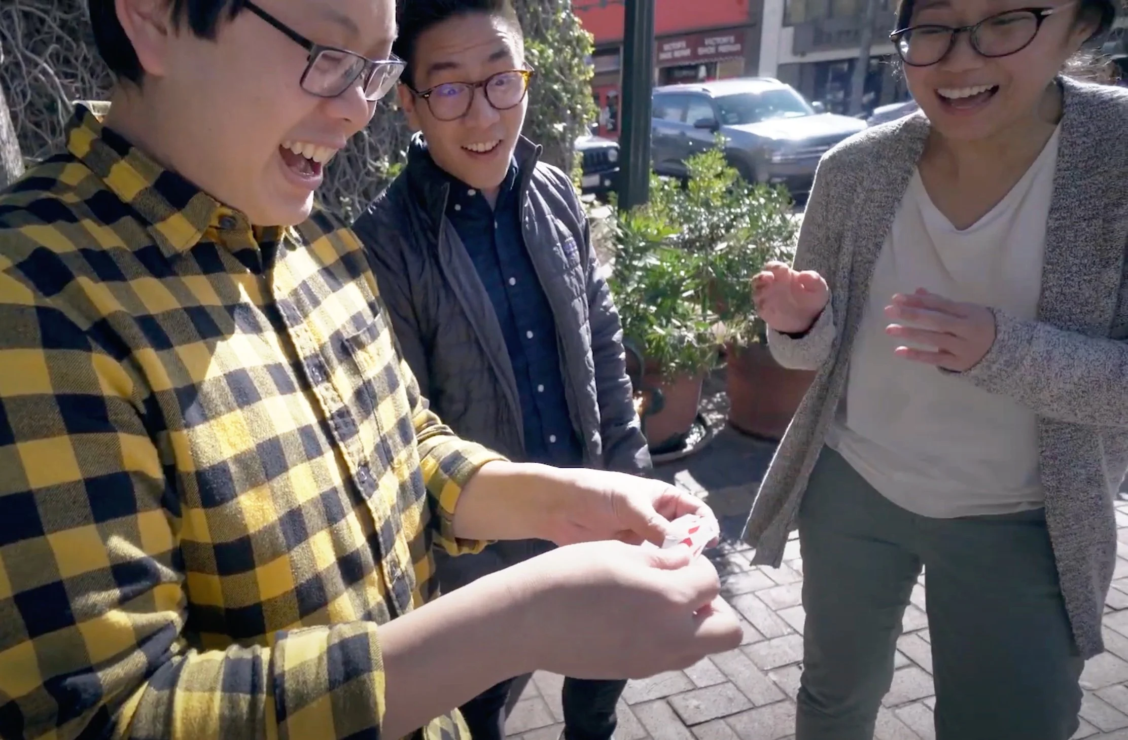 Three people outdoors on a sunny day, laughing and reacting to something one person is holding in their hands. They are dressed casually and appear to be enjoying a lively moment together.