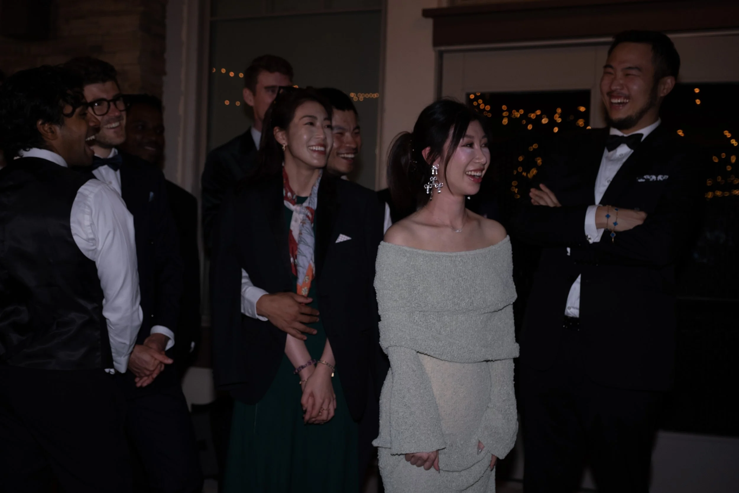 A group of people in formal attire, laughing and smiling, standing indoors at a social event during evening or nighttime.