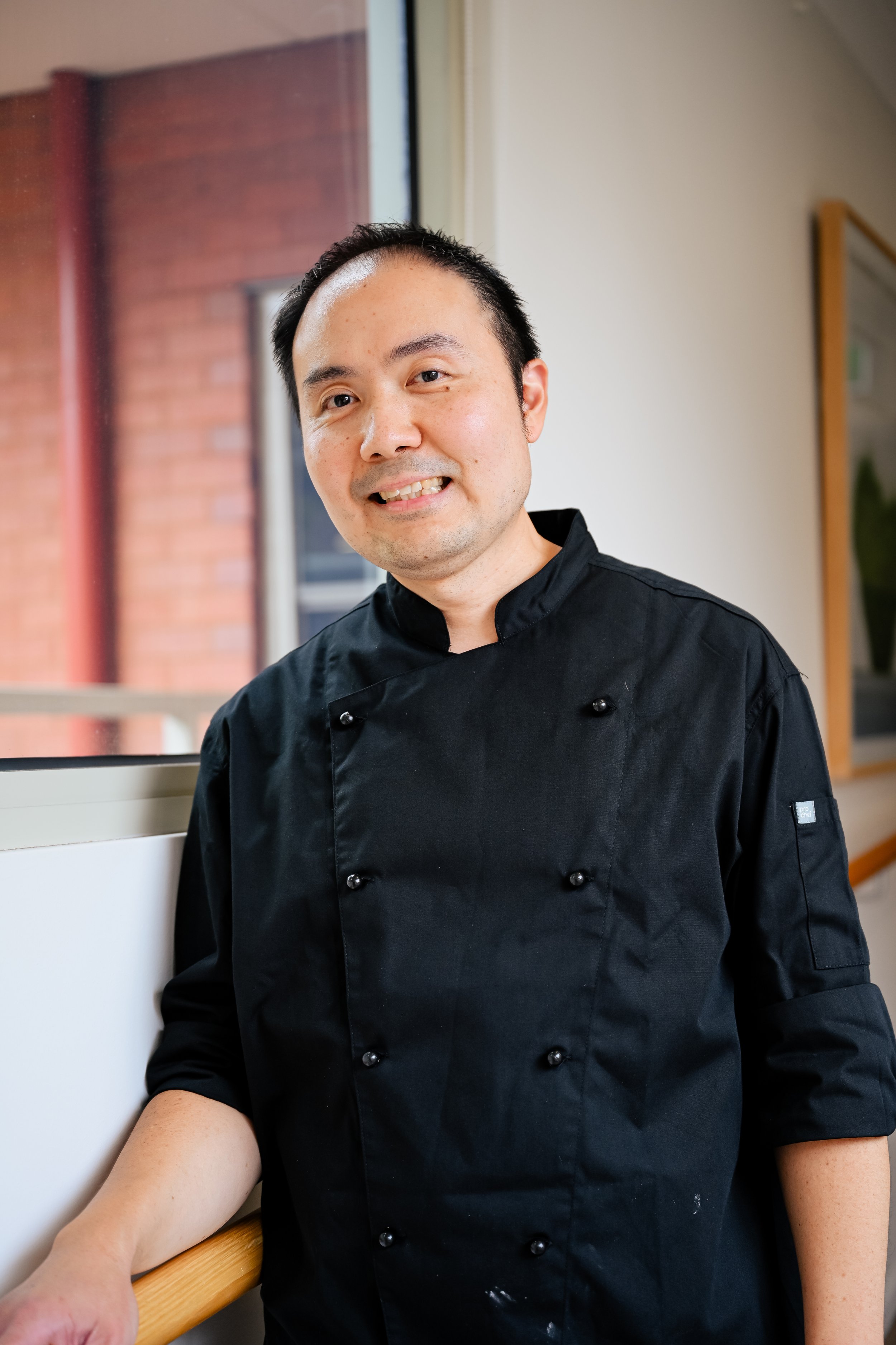 Portrait of a smiling man wearing a black chef's coat, standing in a room with brick and beige walls near a window.