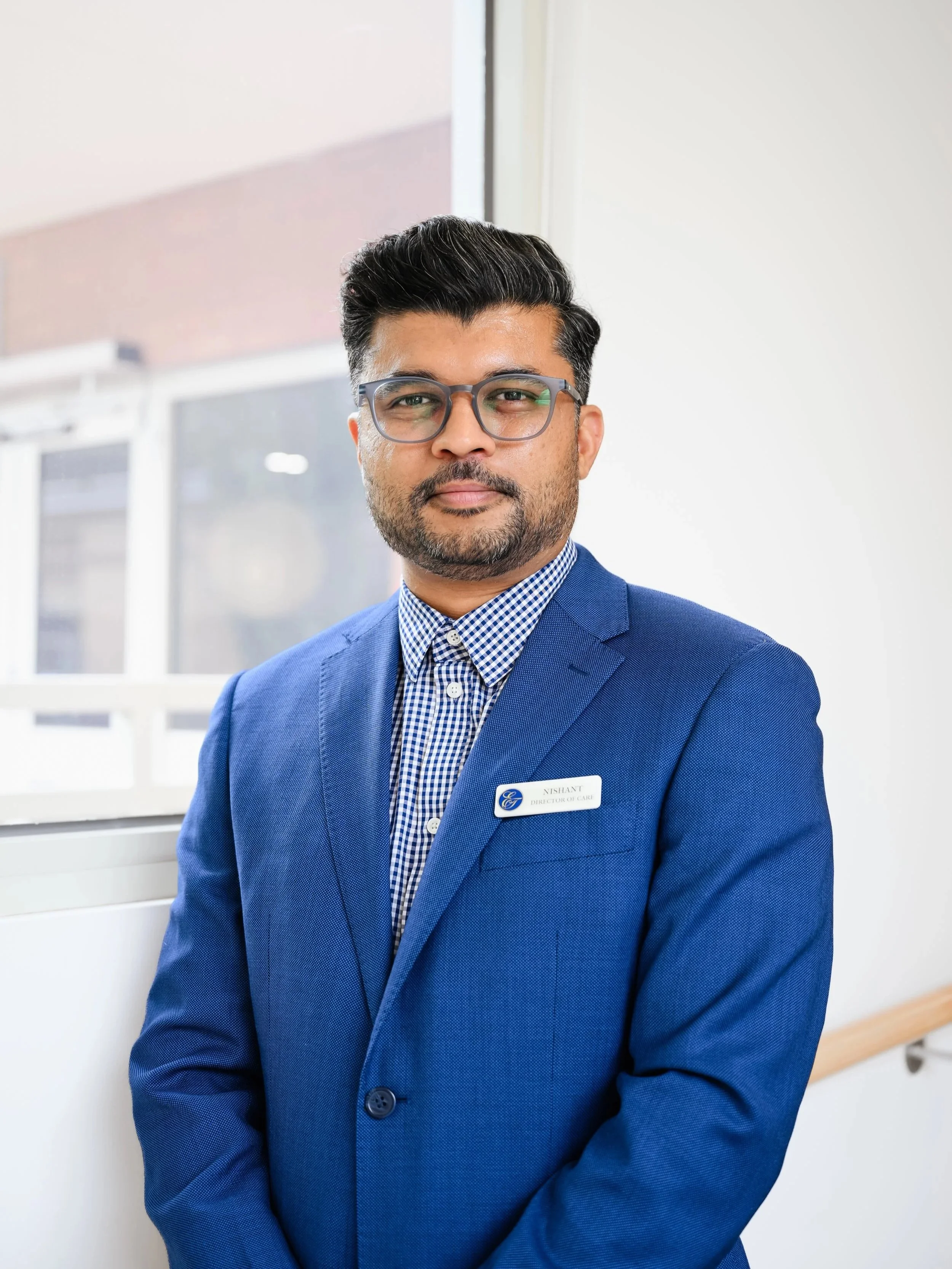 A man in a blue suit with glasses standing indoors near a window, wearing a name badge that reads 'Nishant, Director of Care'.