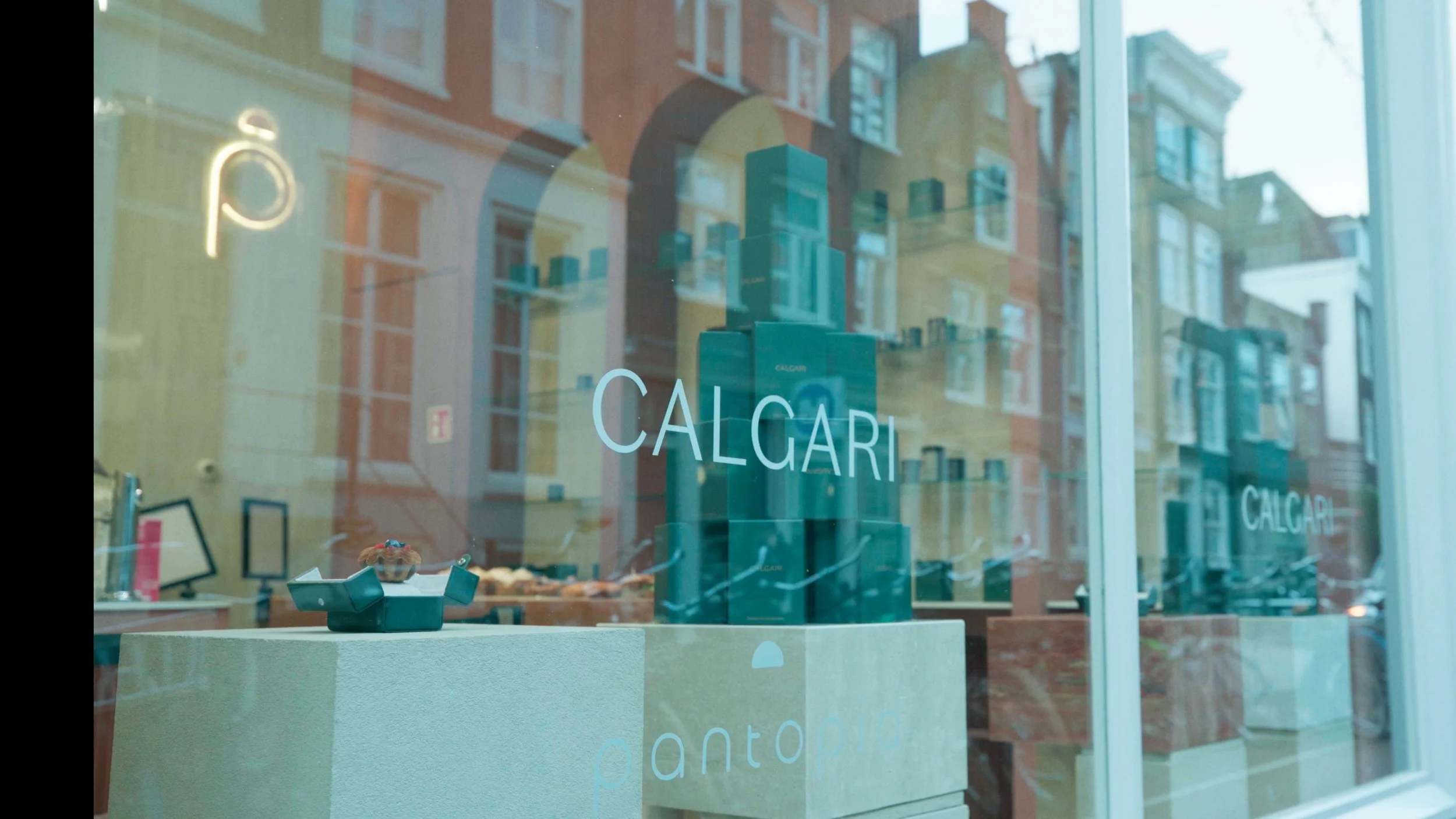 Gezicht op de etalage van een winkel met de naam 'CALGARI'. Men ziet een witte tafel met decoraties en een glazen deur met de winkelnaam erop. De reflectie van de straat met gebouwen is zichtbaar in de ramen.