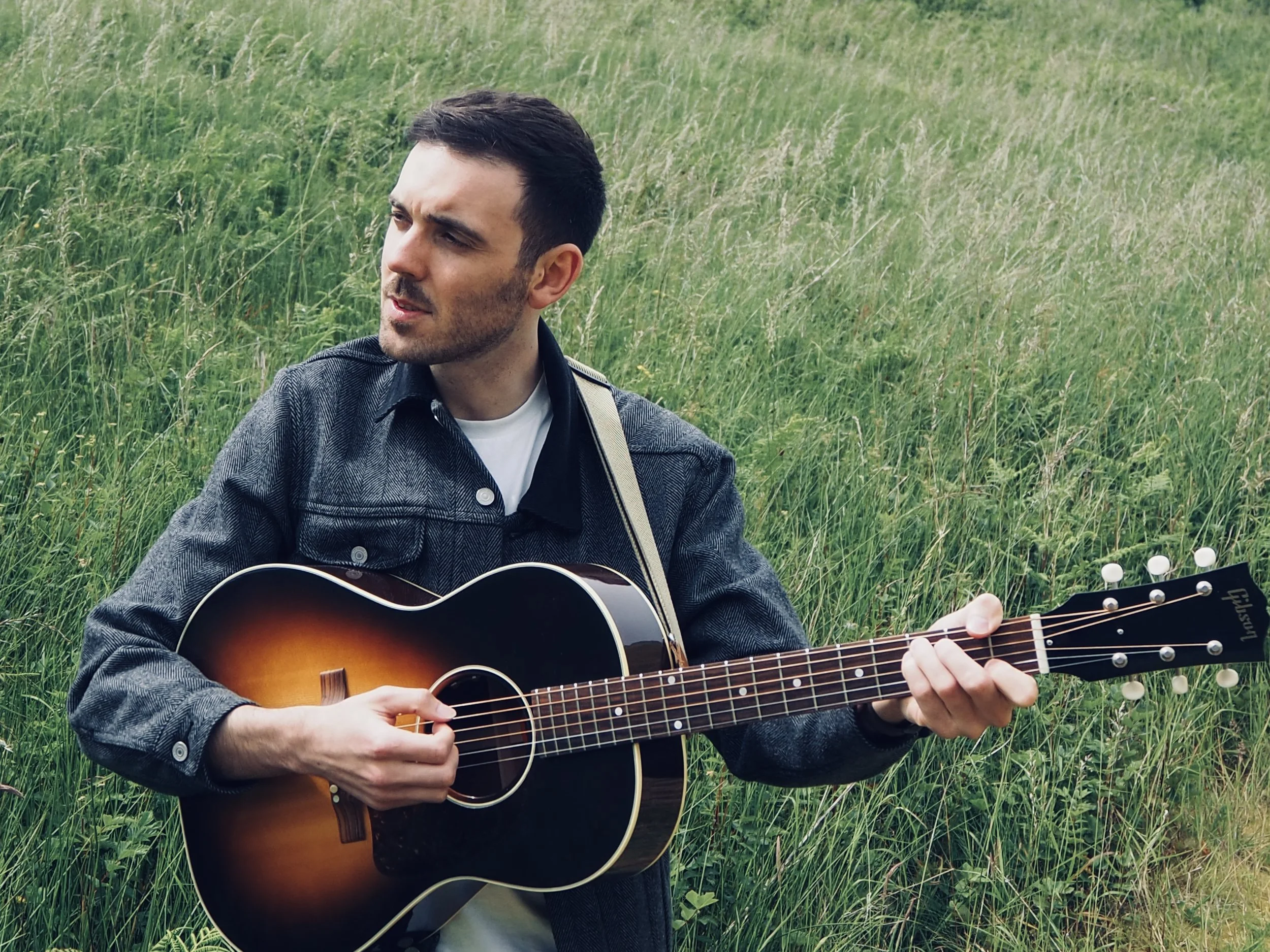 A man playing an acoustic guitar outdoors in a grassy field.