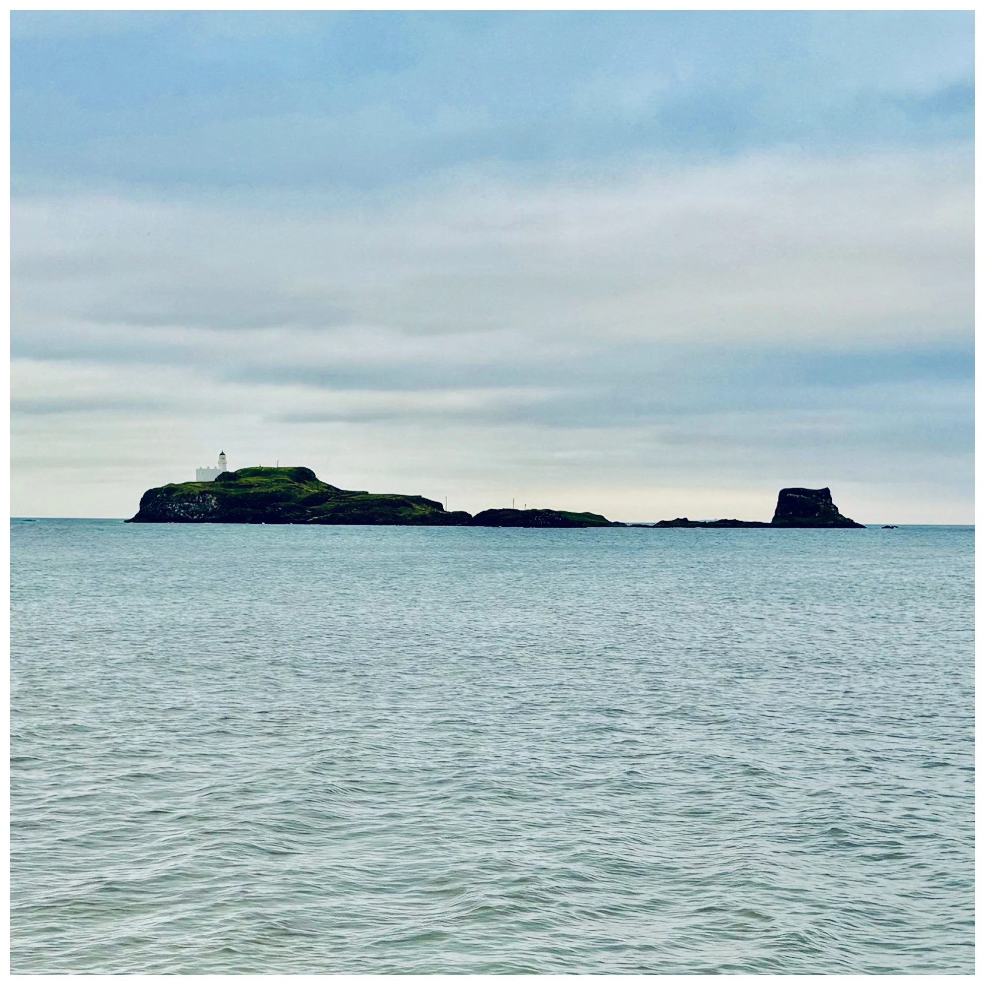 A scenic view of an island with a lighthouse and greenery, surrounded by calm ocean waters under a cloudy sky.