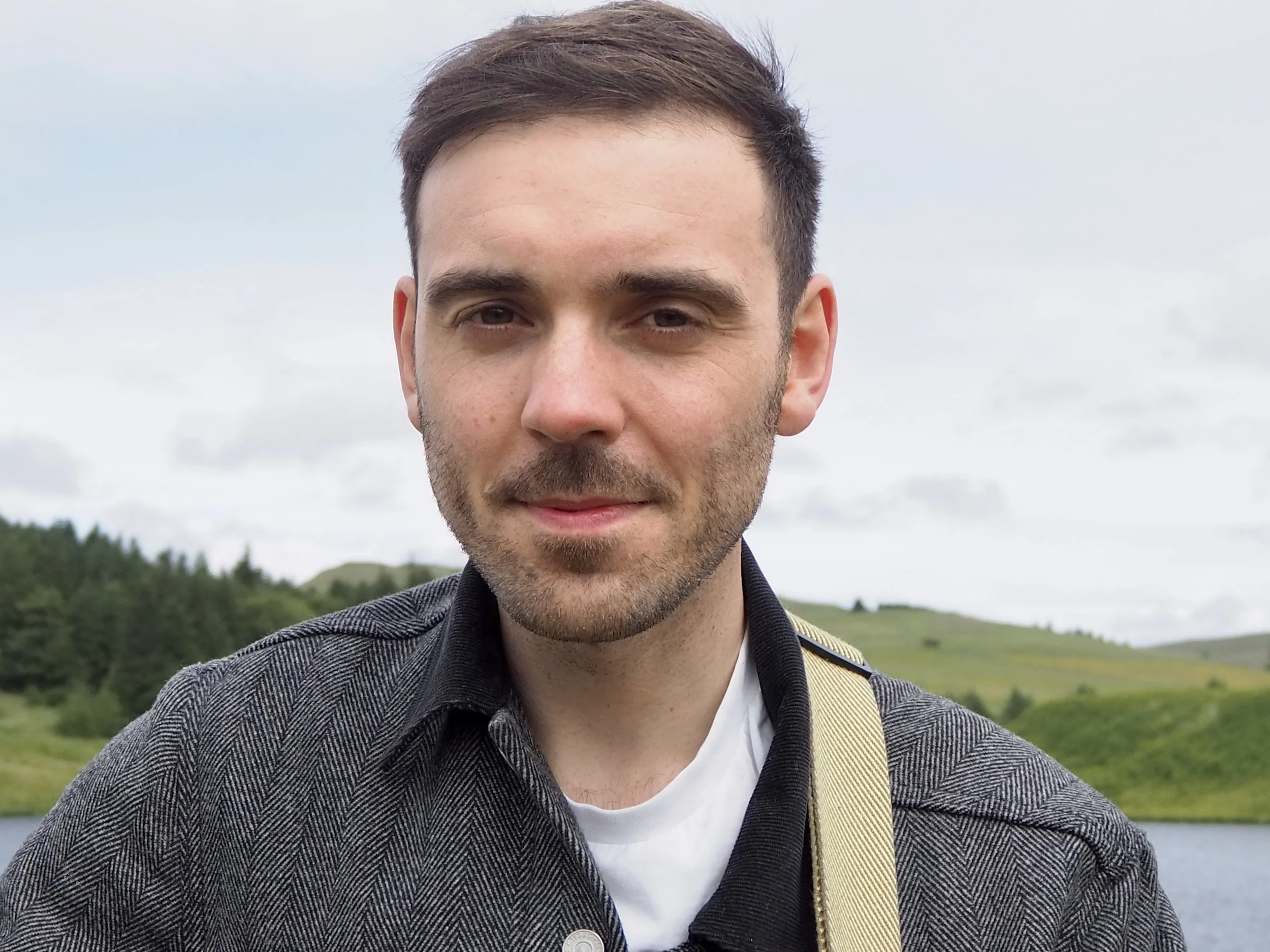 Close-up of a young man with short brown hair and a beard, smiling, outdoors with a lake and green hills in the background.
