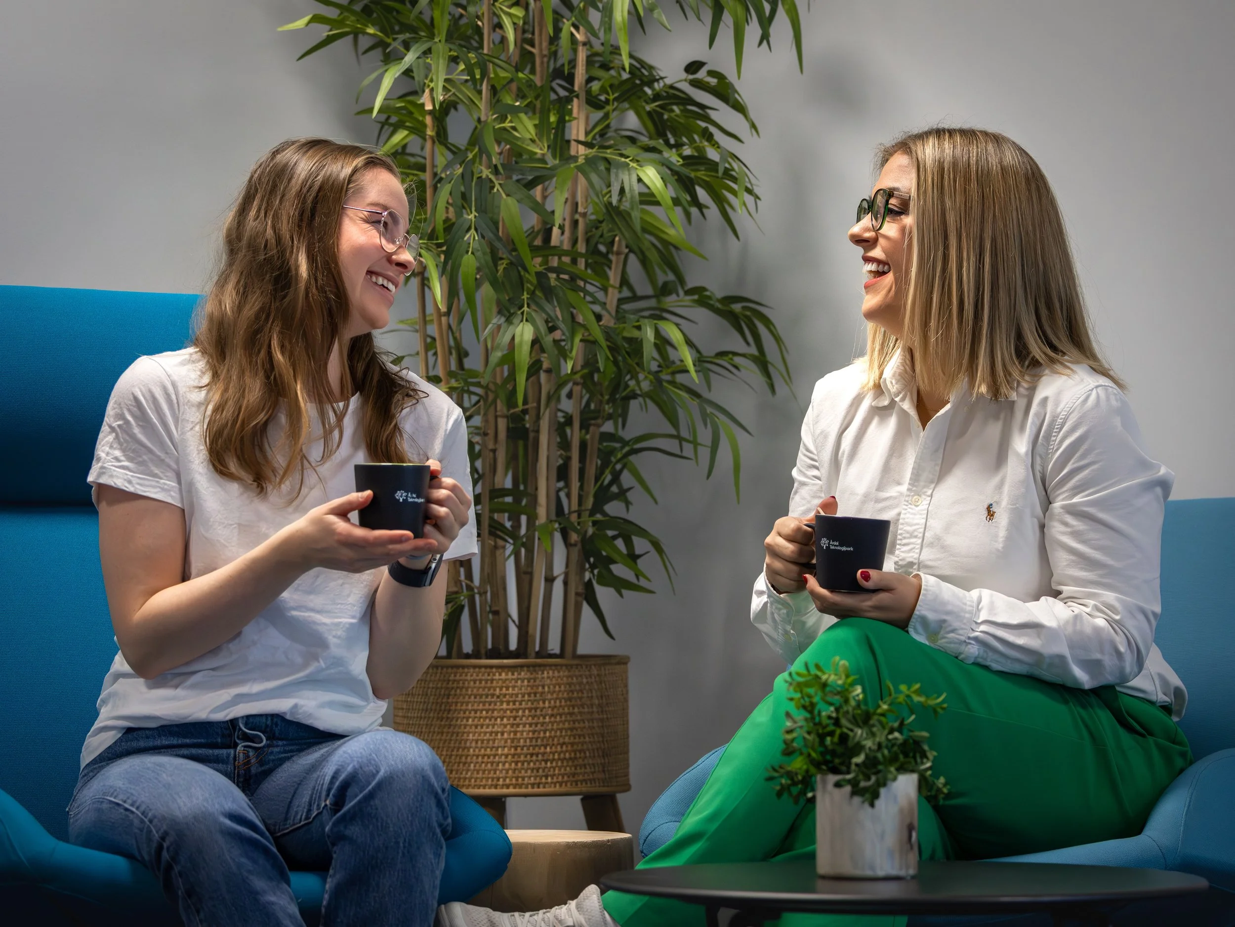 To kvinner som smiler og snakker mens de drikker kaffe i et hyggelig uformelt miljø med planter og blå sofaer.