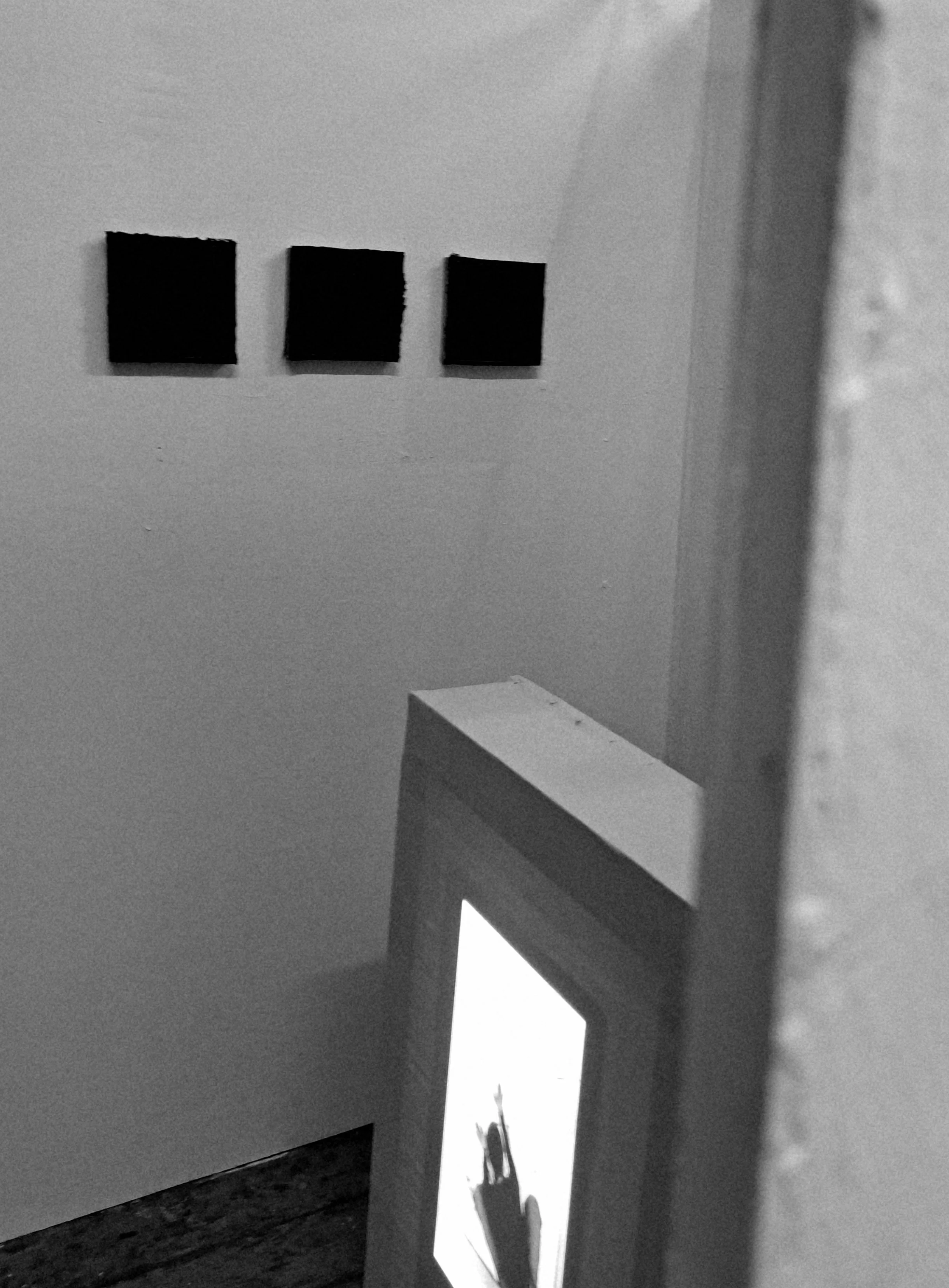 Black and white photo showing an art instillation, containing a collection of three art pieces hung on a wall, with a freestanding canvas being projected onto partially visible in the foreground.