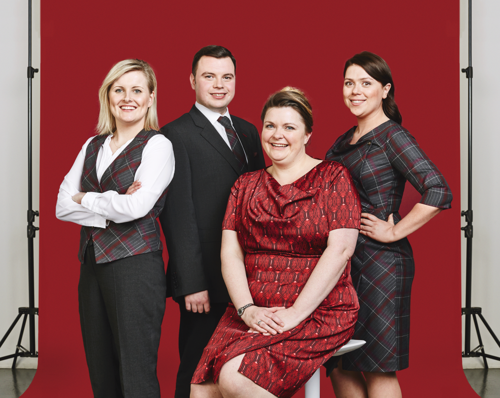 Four smiling adults, three women and one man, posing in front of a red backdrop during a photo shoot. The woman in the center is seated, wearing a red dress, while the other three stand around her.