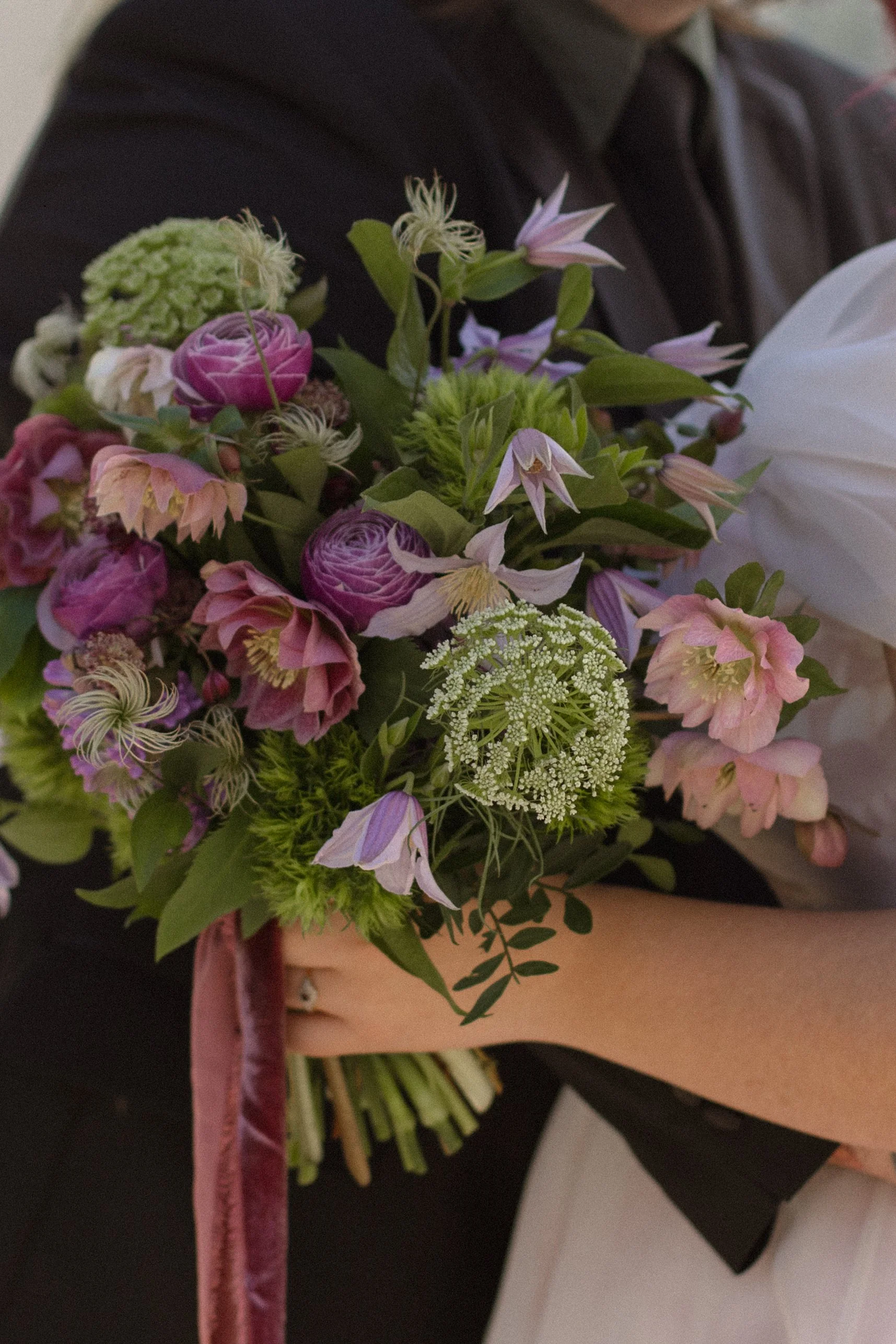 A springtime bridal bouquet featuring ruffled pink hellebores, whimsical lilac clematis and its fluffy seedheads, frothy ammi and other blooms in shades of green, purple and pink.
