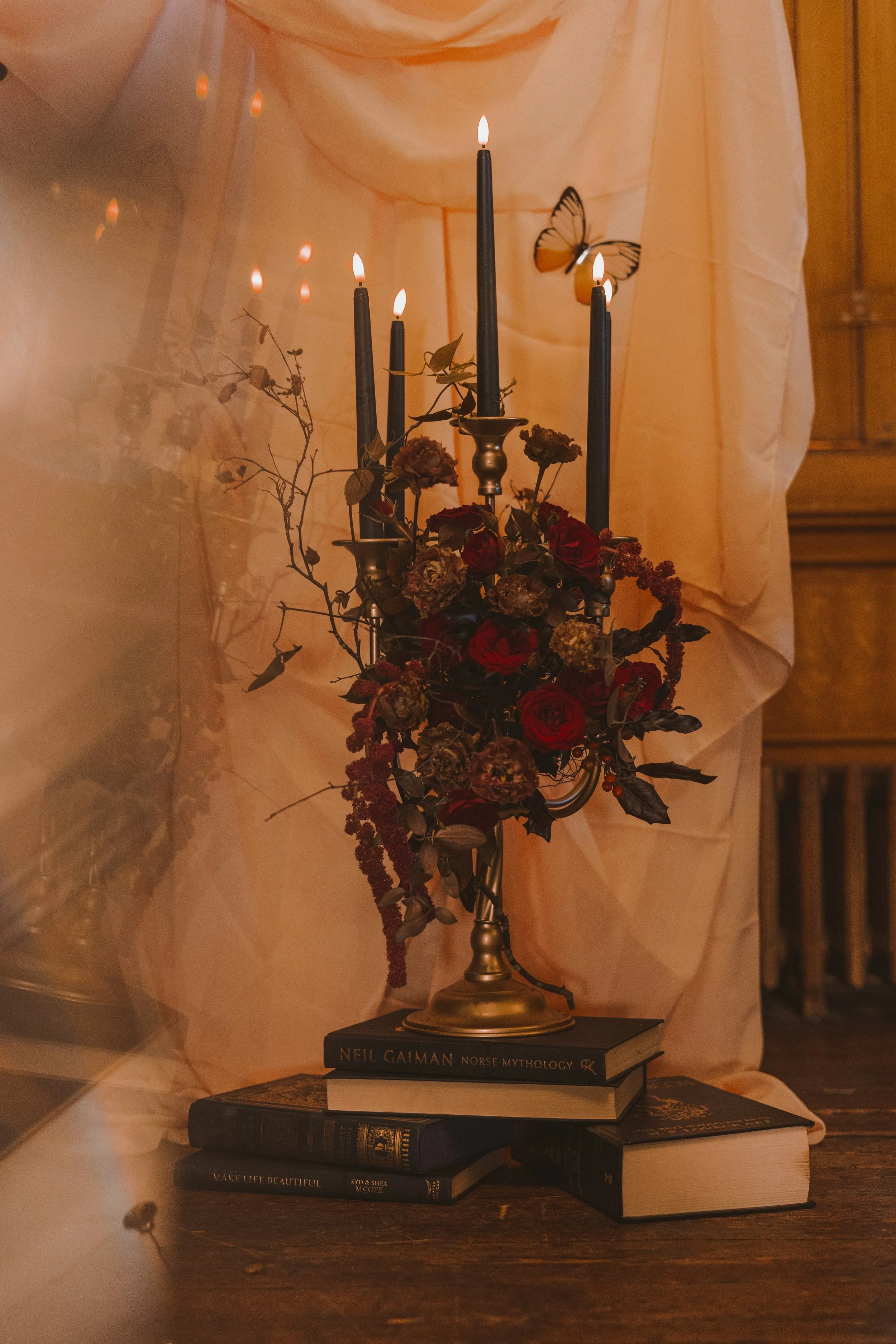 An antique bronze candelabra with black candles adorned with moody, autumnal flowers like trailing amaranthus, brown lisianthus and red roses. The candelabra is stood on a pile of vintage books in front of a draped pale pink background