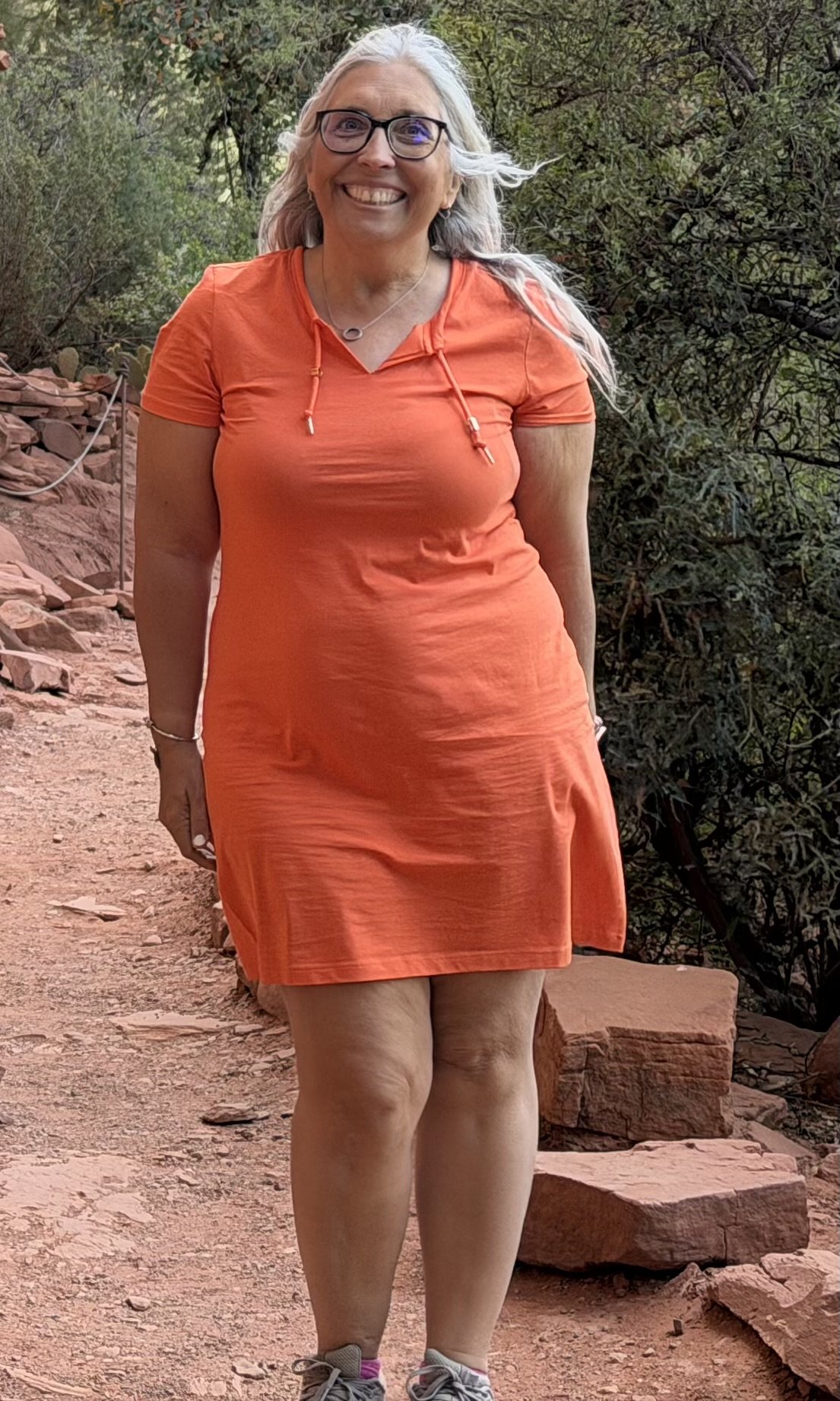 Woman with gray hair wearing glasses and an orange dress, standing on a rocky dirt trail with trees in the background.
