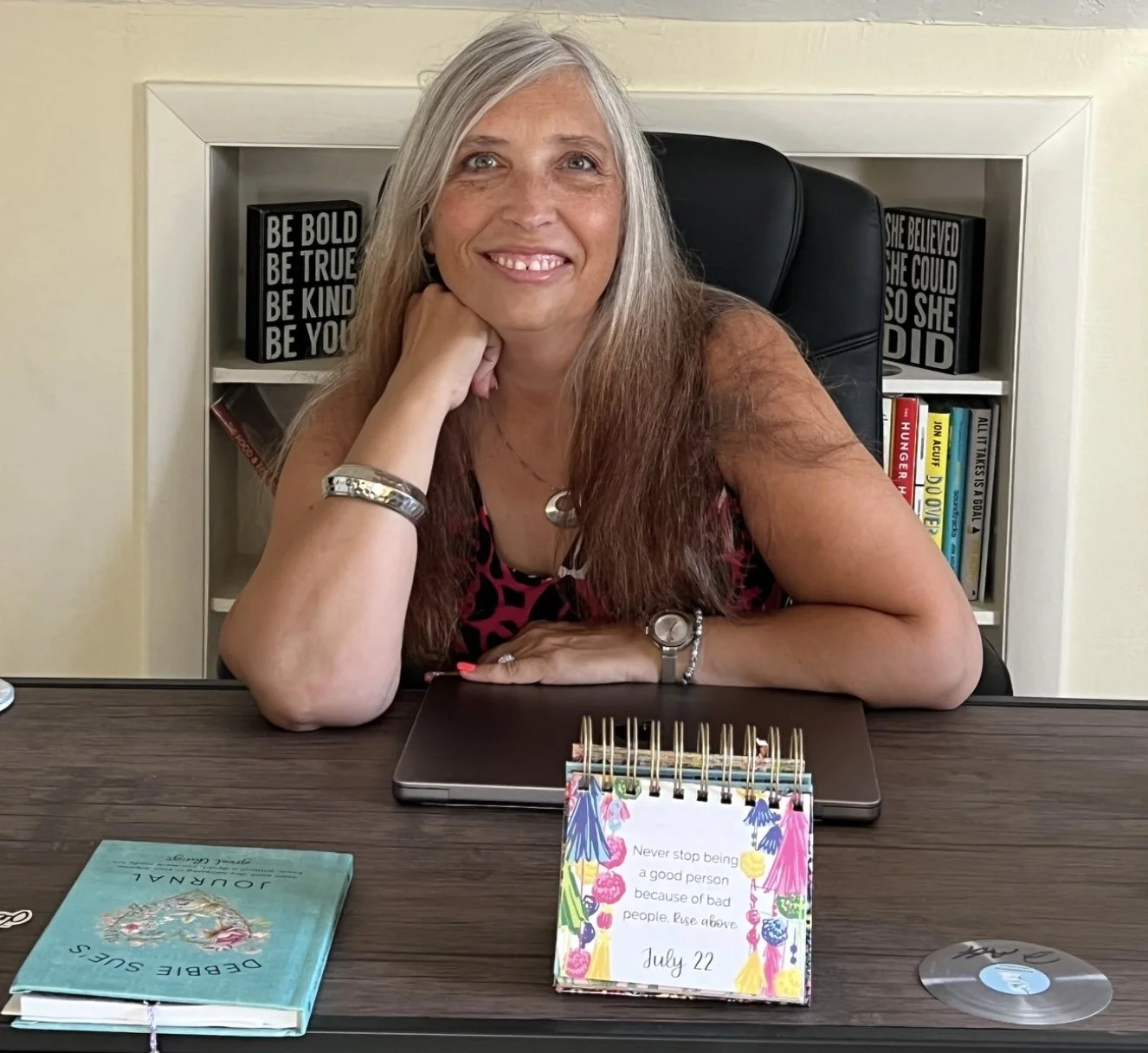 A woman with long gray hair, smiling, sitting at a desk with a book, a colorful calendar that says July 22 and motivational quote, and a laptop, in an office with bookshelf and motivational signs in the background.