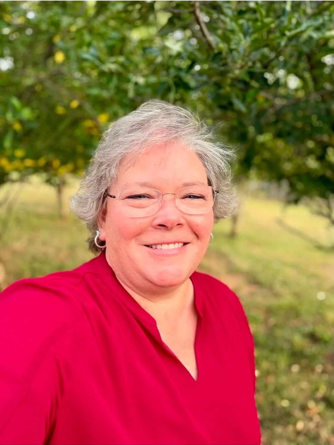 A smiling woman with short gray hair, glasses, and hoop earrings, wearing a red top, standing outdoors in front of green trees.