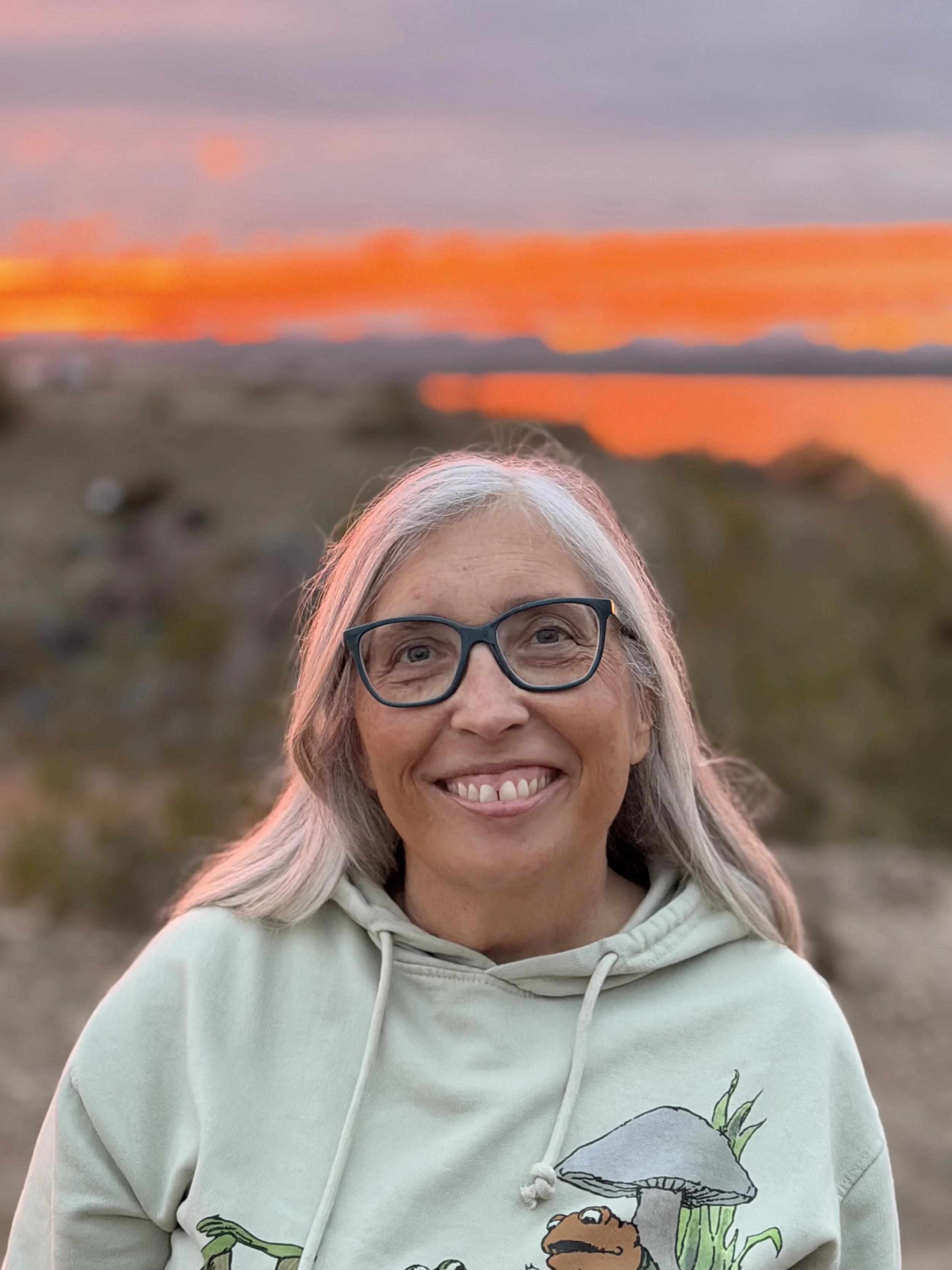 A smiling woman with gray hair and glasses wearing a light-colored hoodie with a mushroom and fairy illustration on it, outdoors during sunset with a blurred background of rocks and water reflecting the orange and purple sunset colors.