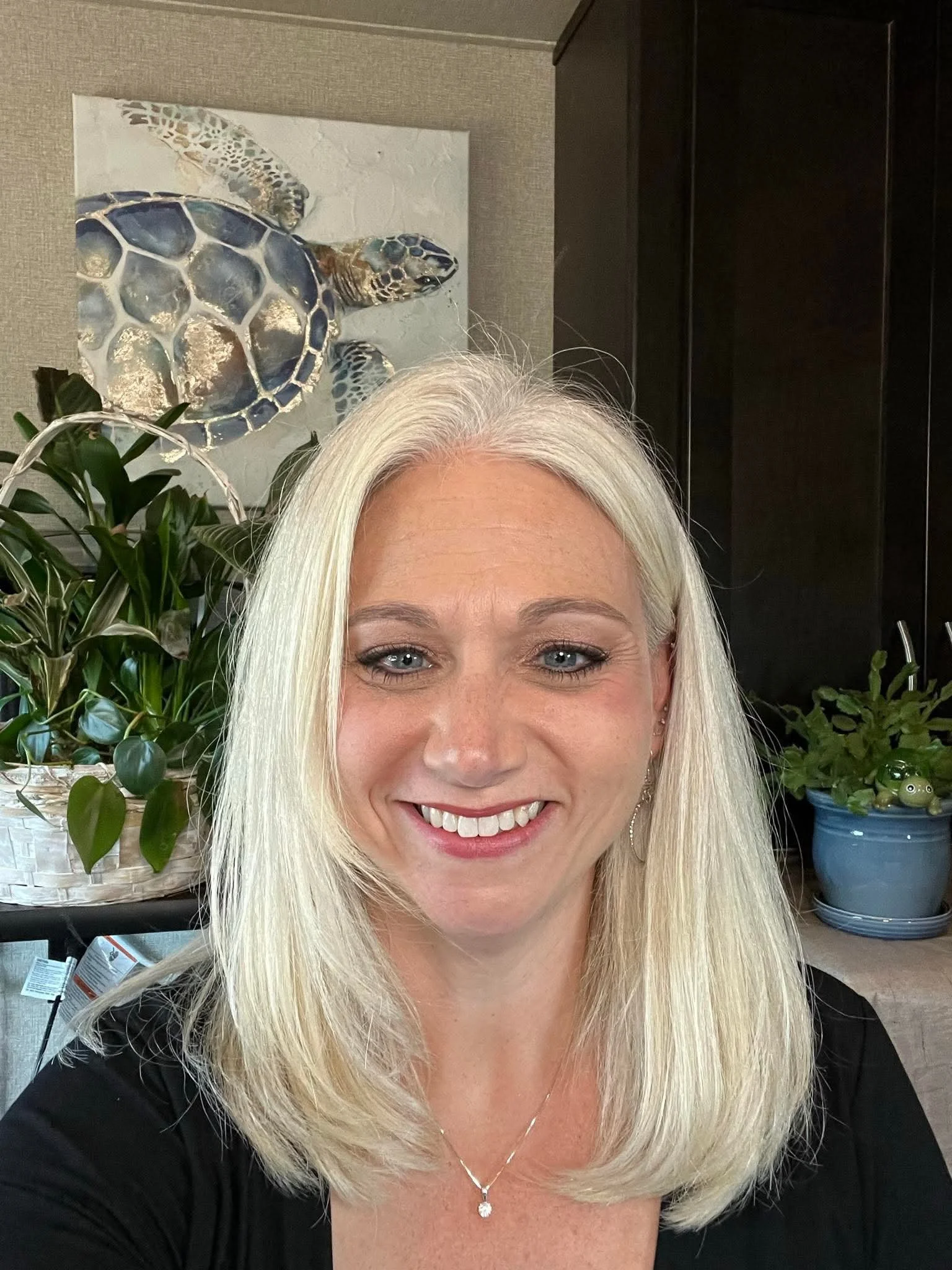 A smiling woman with long blonde hair and blue eyes takes a selfie indoors. Behind her, there is a painting of sea turtles on the wall, green plants in baskets, and a dark piece of furniture.