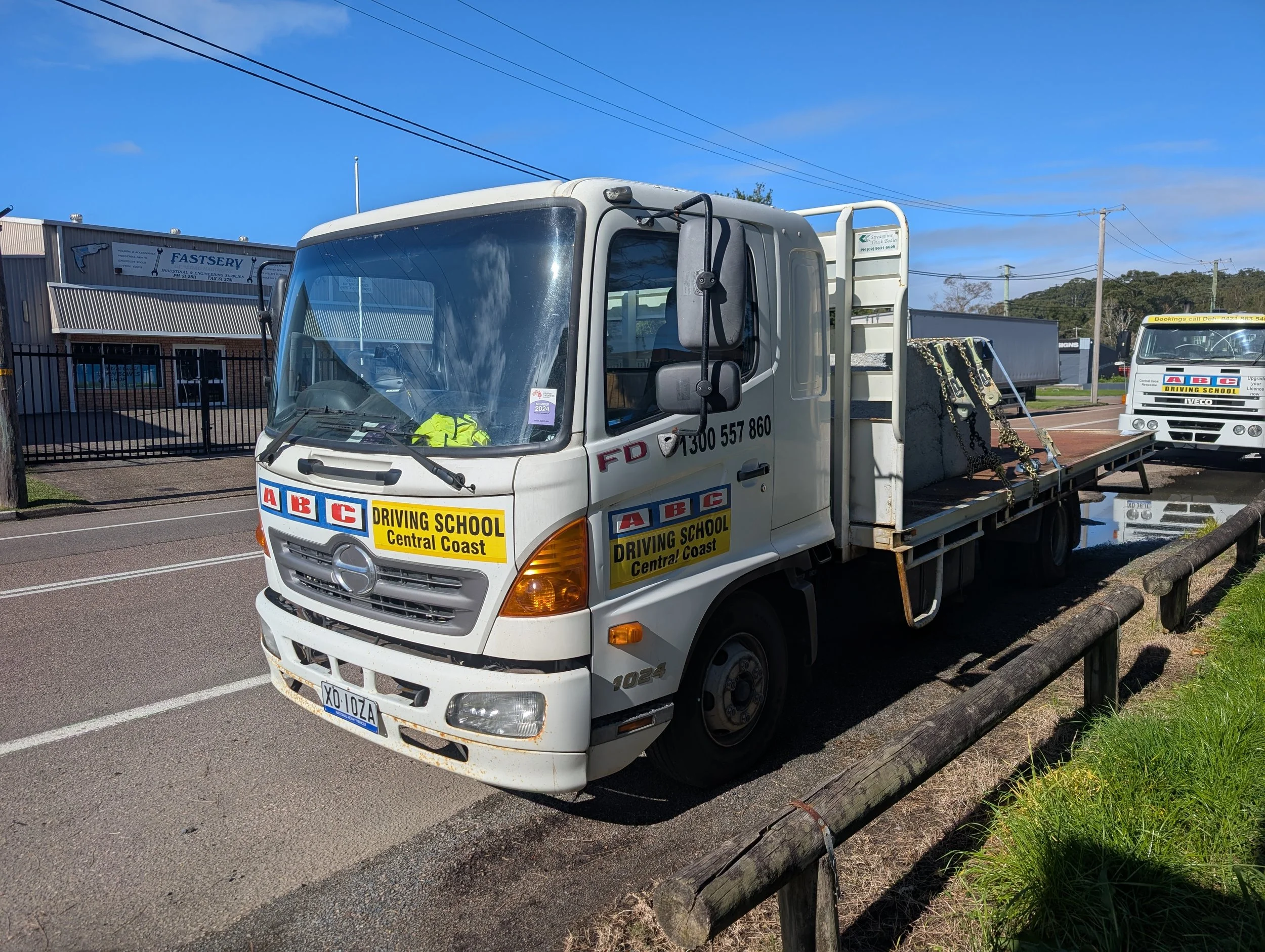A white flatbed truck with signs for ABC Driving School Central Coast, parked on a street with a supermarket in the background and power lines overhead.