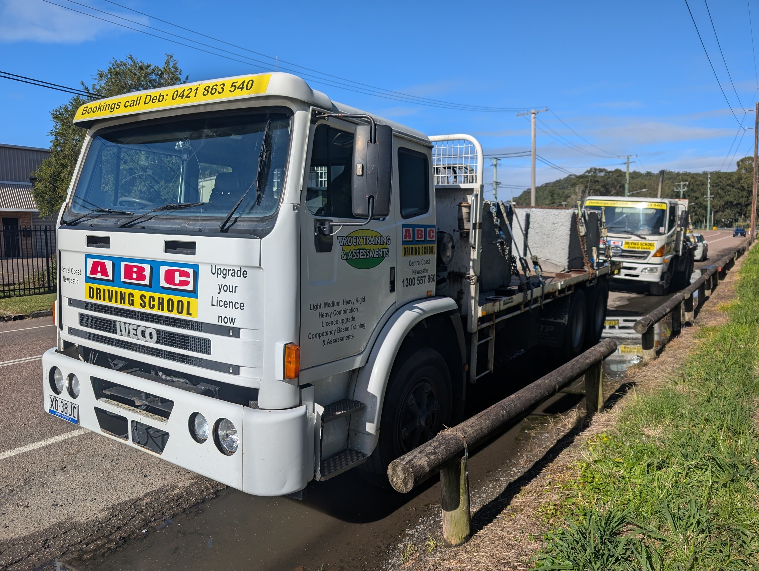 White flatbed truck parked along the side of the road with signs advertising a driving school, ABC Driving School, and contact information for bookings. The truck is next to a wooden post barrier and there are suburban houses and power lines in the background. The sky is clear and blue.