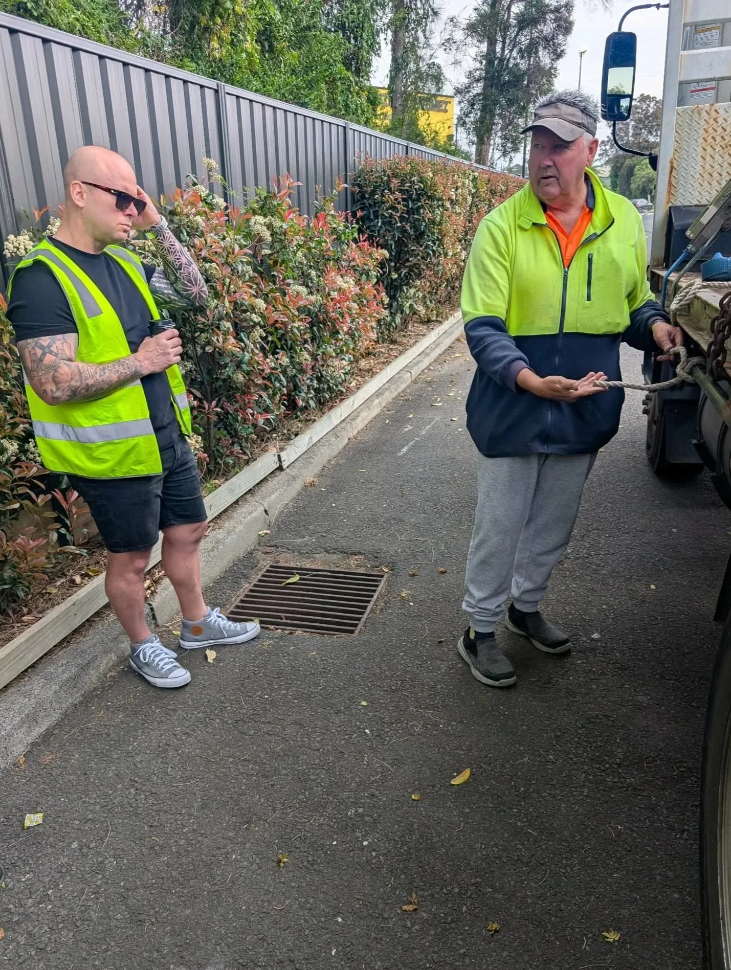 Two men standing beside a truck on a paved street, engaged in a conversation. One man is wearing a lime green and black jacket, gray sweatpants, and a cap, holding a rope. The other man is dressed in a black T-shirt, dark shorts, gray sneakers, sunglasses, and has tattoos and a tattooed arm, holding a cup.