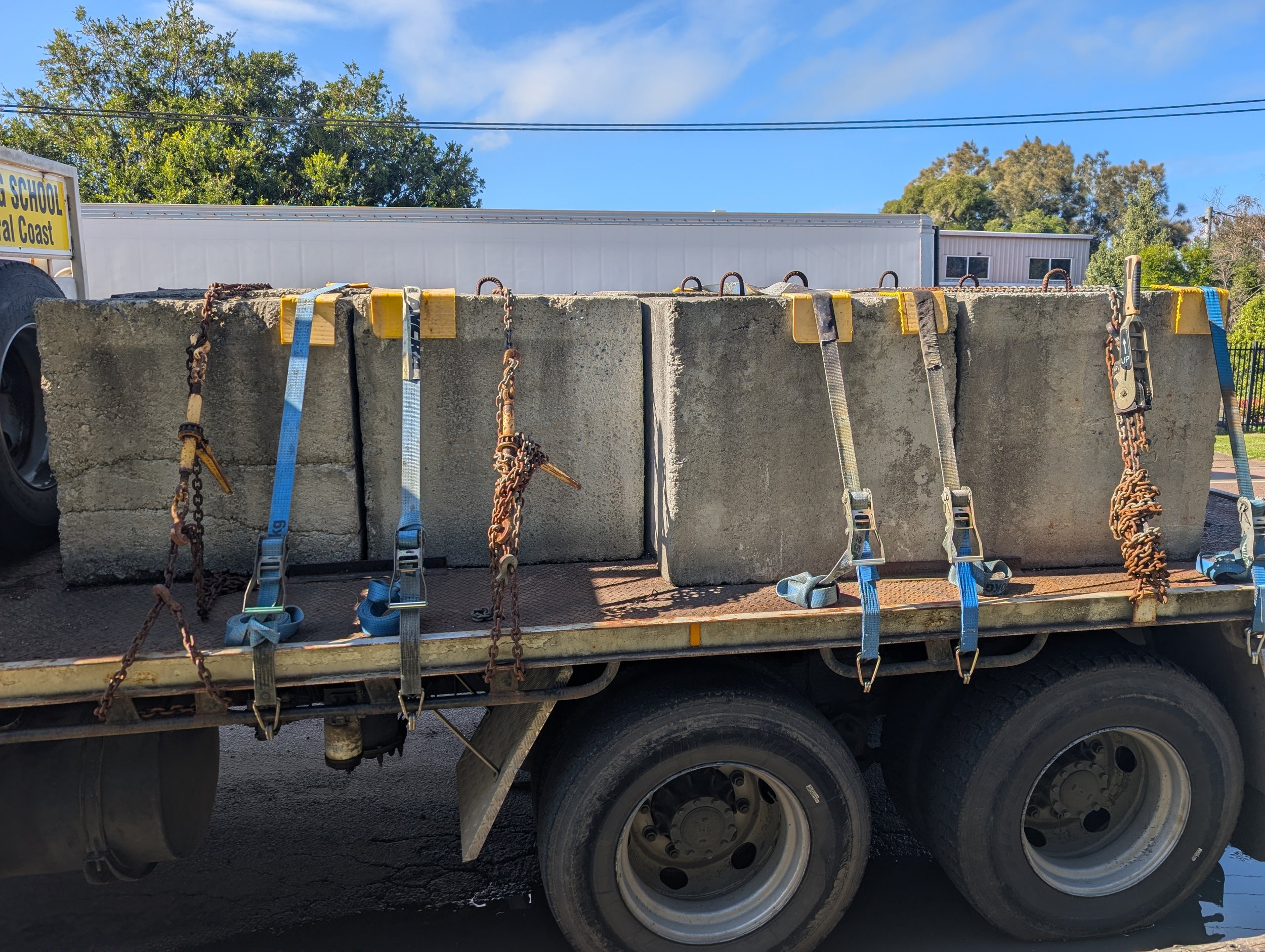 Concrete blocks secured with straps and chains on a flatbed truck, with trees and a building in the background.