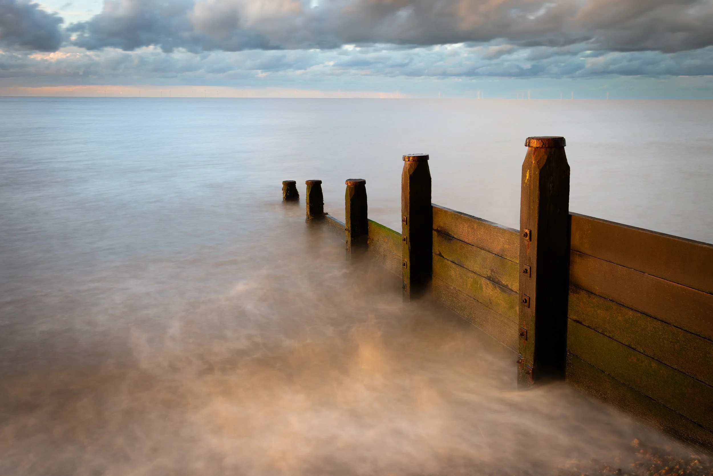 A weathered wooden sea barrier extending into a calm ocean, with a cloudy sky overhead.