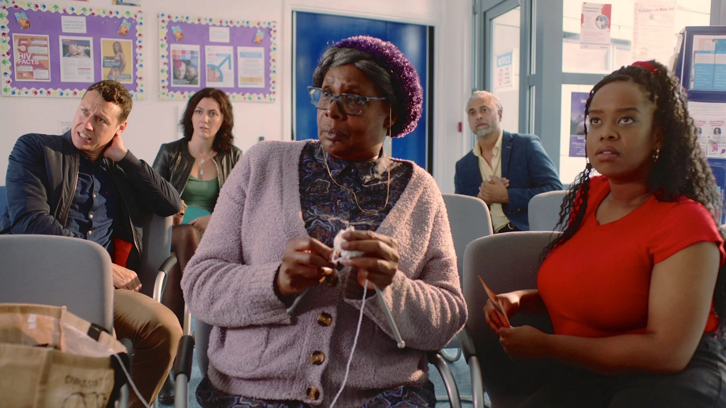 A diverse group of five people sitting in a waiting room or classroom, appearing attentive. The woman in the foreground wears glasses, a pink cardigan, and a patterned dress, holding a tissue. The young girl next to her is in a red shirt, looking upward. The other three individuals in the background have varied expressions, seated near bulletin boards and windows.