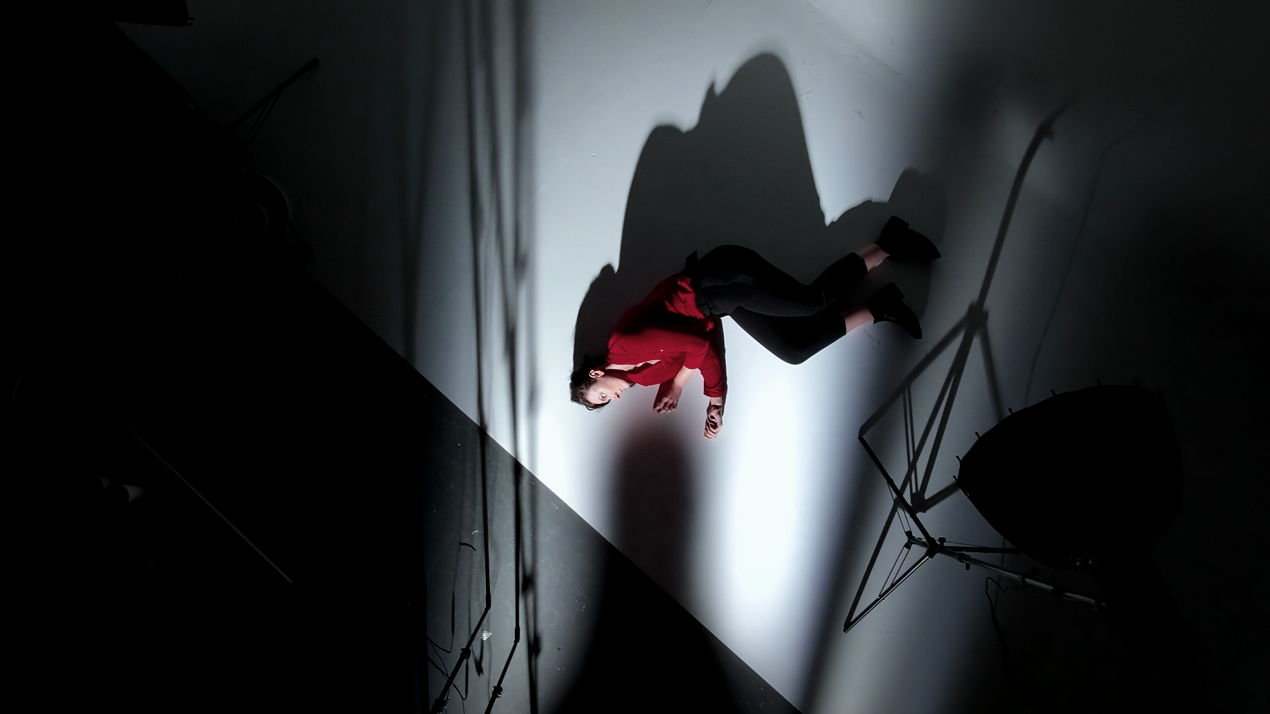 A person dressed in a red jacket and black pants, crouching on the floor next to a projector and a tripod, with dramatic lighting creating large shadows on the wall.