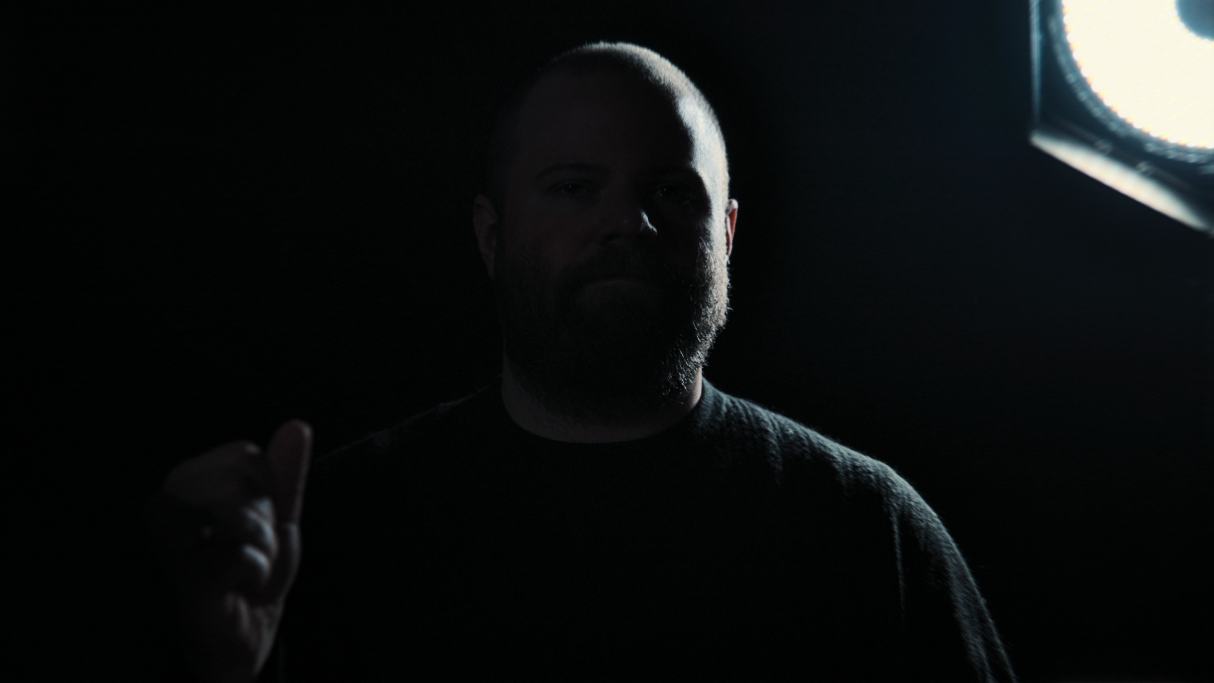 A man with a beard and a bald head stands in a dark environment with a bright light source to the right, partially illuminating his face and hand.