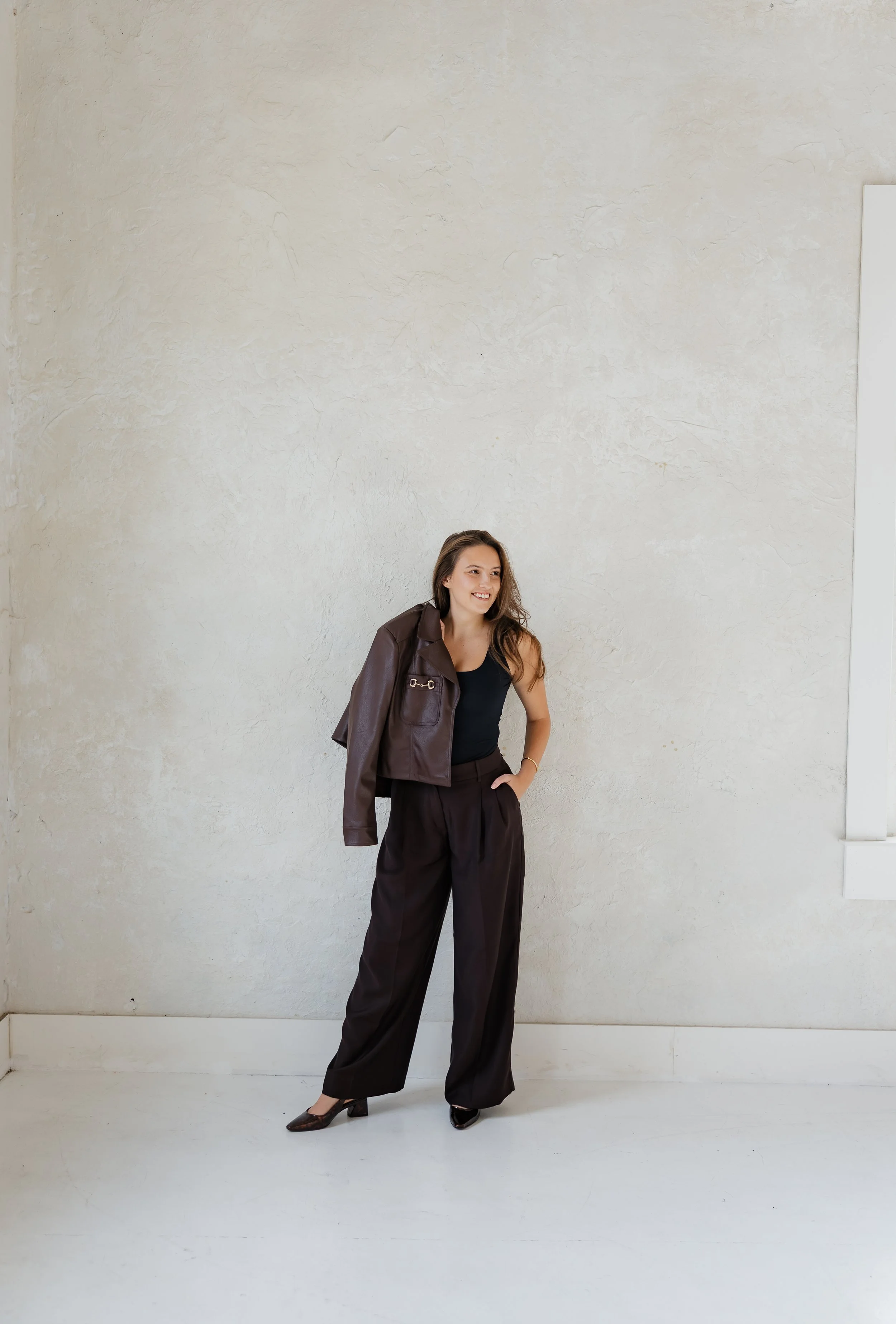 Woman standing in a fashion pose against a minimalist, textured off-white wall, smiling and holding a brown leather jacket over her shoulder, wearing a black top and wide-leg pants with heels.