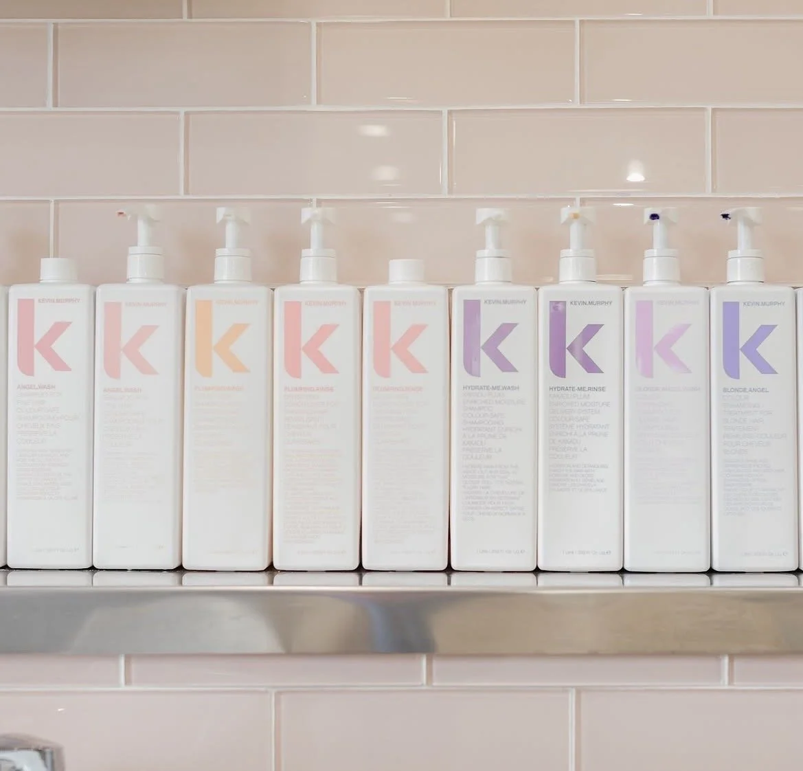 A row of nine white bottles with pump dispensers, labeled with pink, orange, purple, and lavender lettering, placed on a metal shelf against a pink tiled wall.