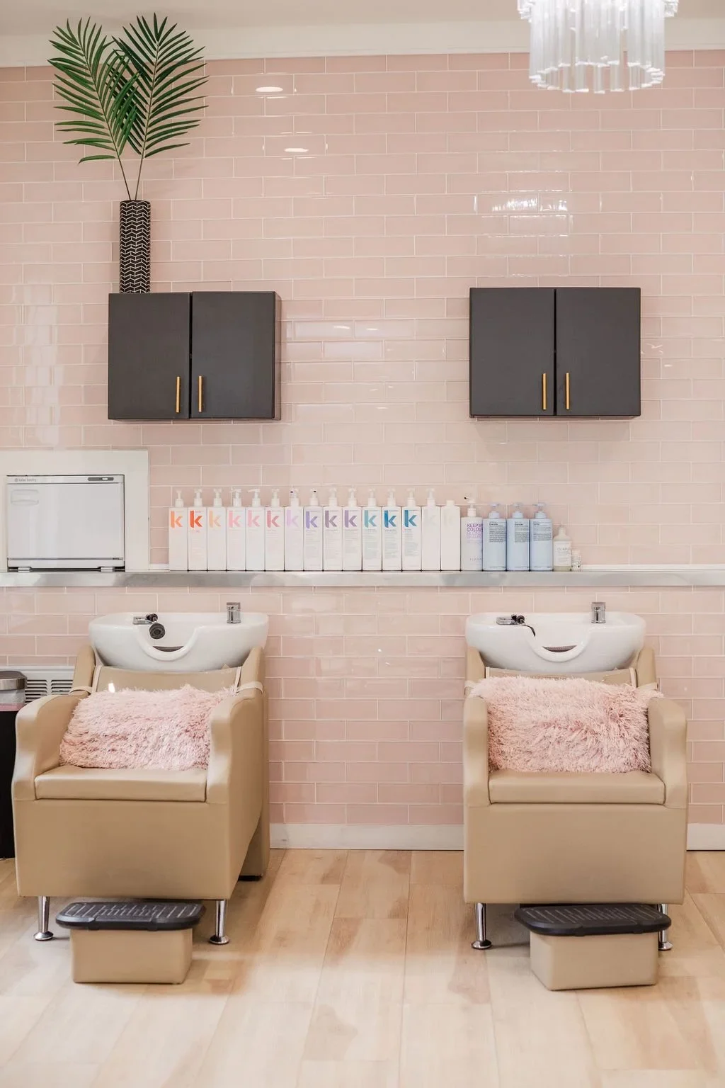 Two beige chairs with pink fuzzy pillows and white sinks inside a salon or spa with a pink tiled wall, black cabinets, a potted plant, and hair care products.
