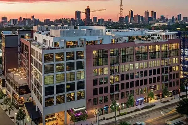 A city street at dusk with a multi-story modern building illuminated from within, and a city skyline with skyscrapers in the background.