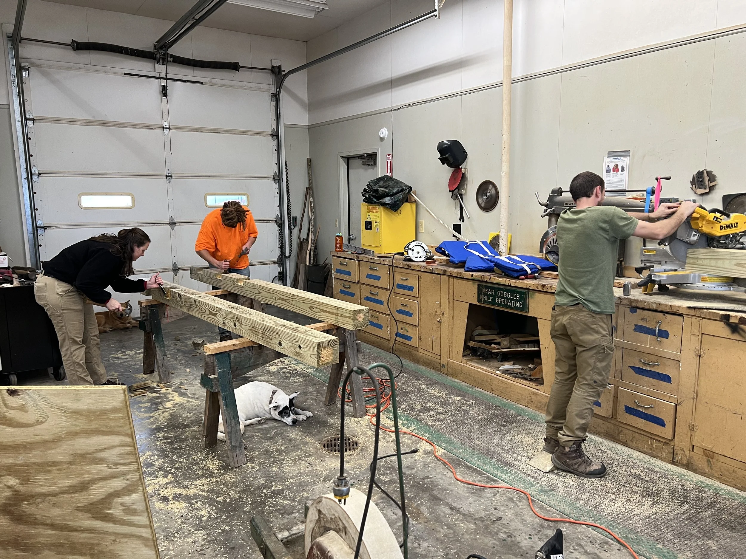 Three people working in a woodworking shop, measuring and cutting wooden beams. A dog is lying on the floor near the workbench, and the workshop has various tools and equipment along the walls.