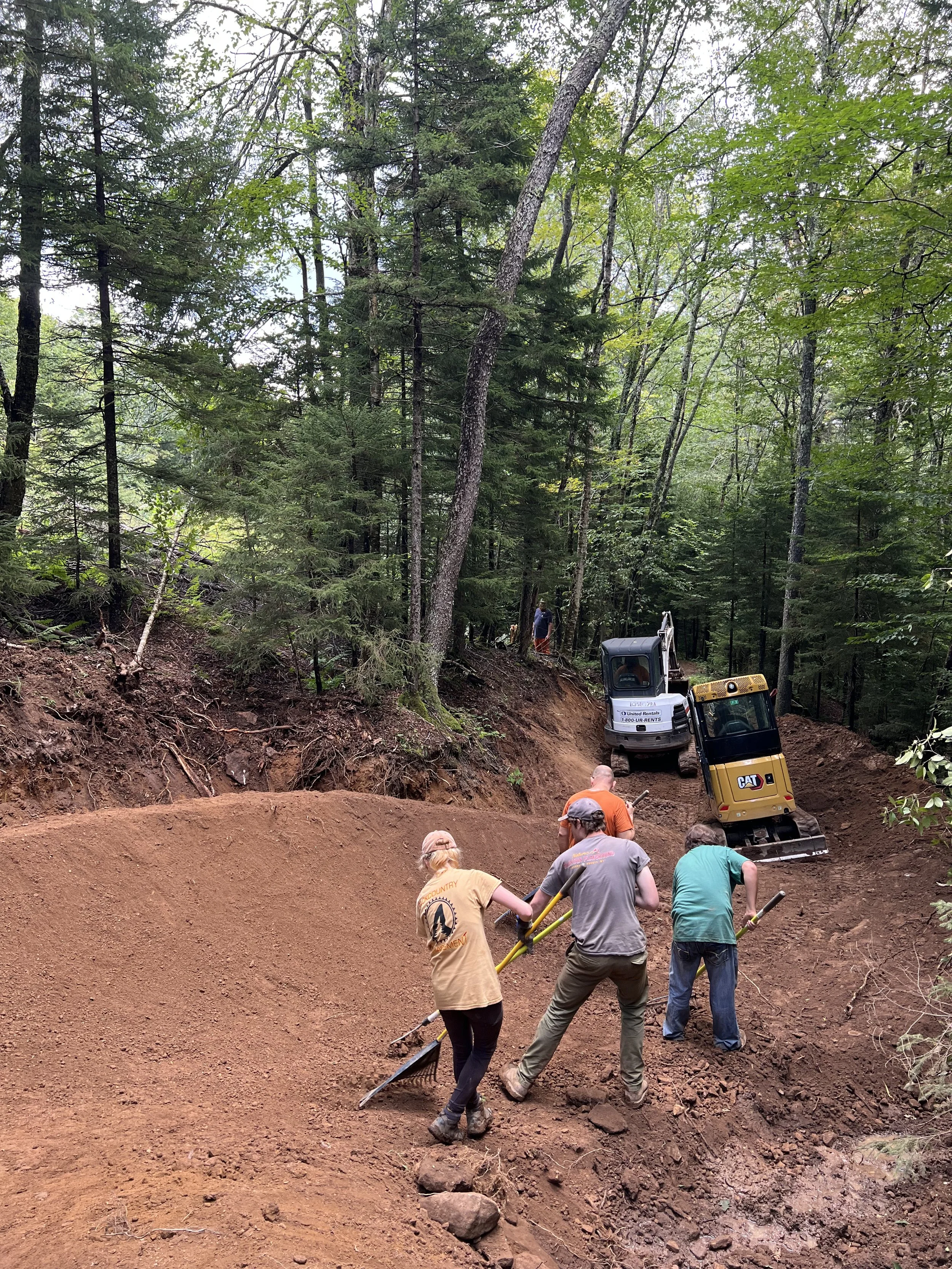 Construction workers leveling dirt on a slope in a forested area with trees and heavy machinery including a small excavator and a skid steer loader.