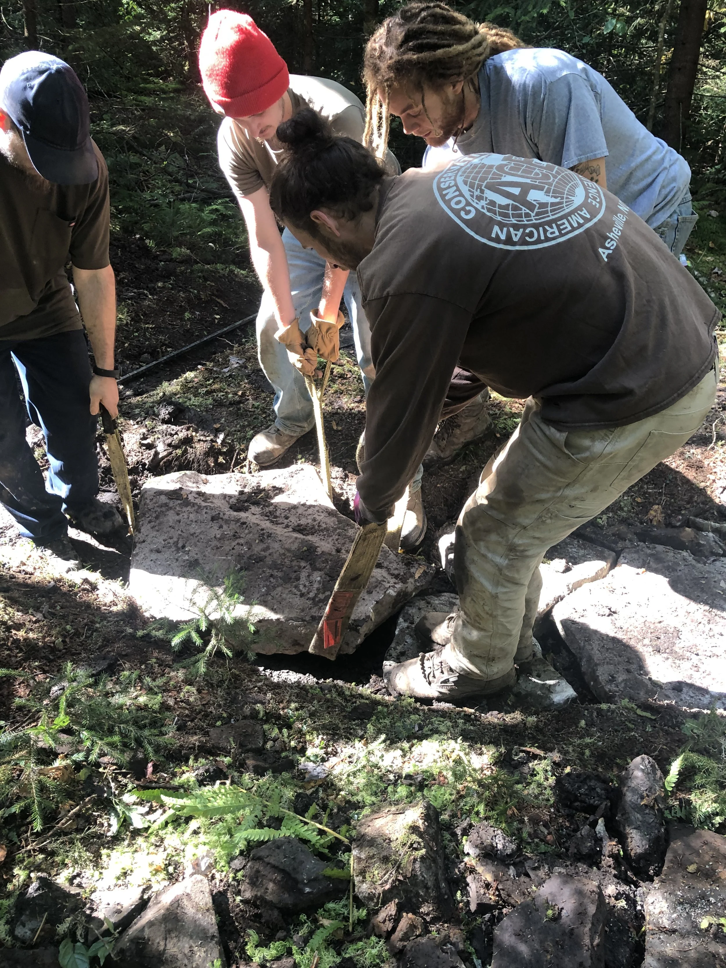 Group of people working together outdoors, lifting or moving a large rock in a forest setting.