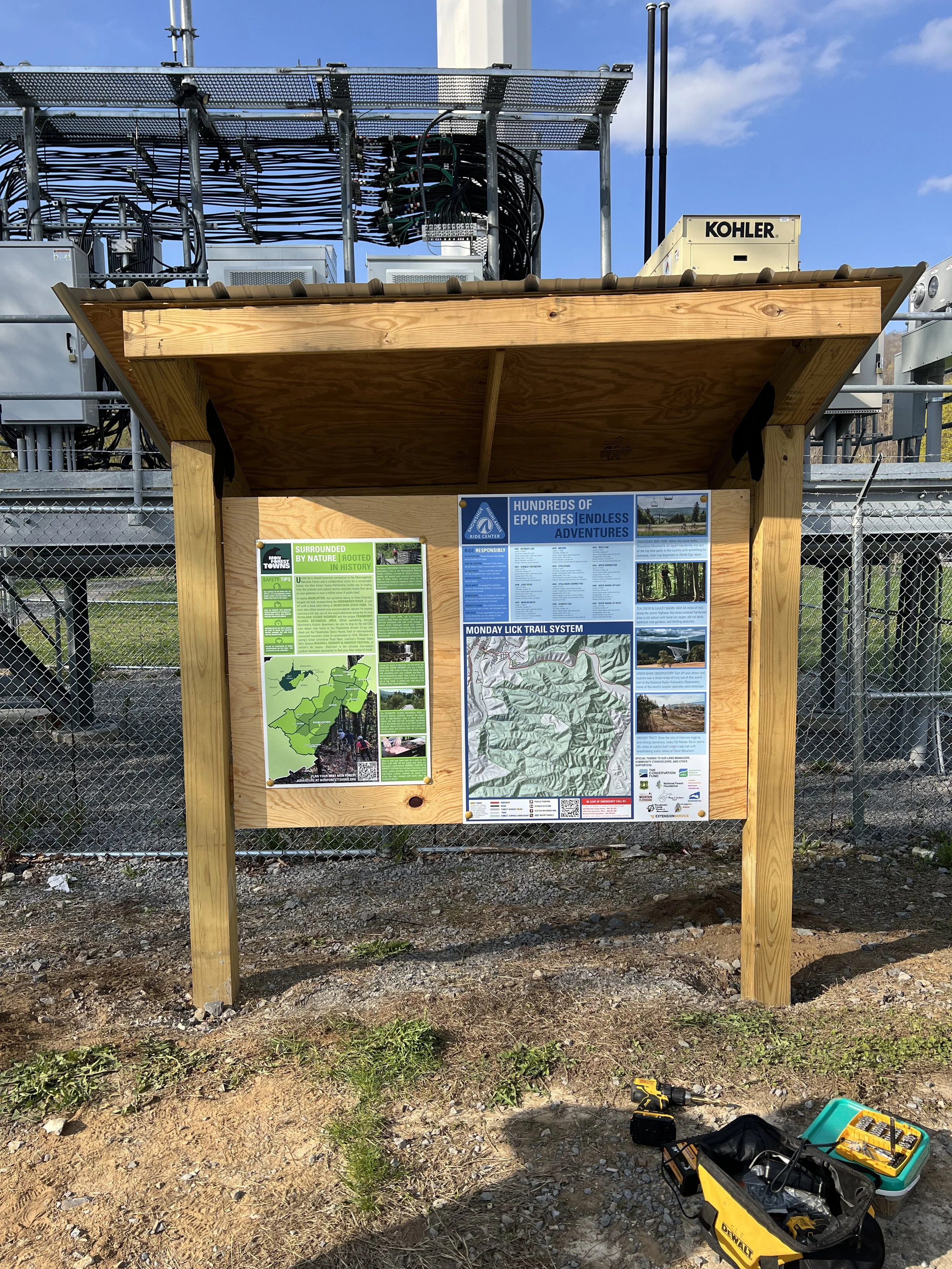 Information board about hiking trails at a forest recreation area, with a map and descriptions, mounted on a wooden stand outdoors, with electrical infrastructure and tools on the ground.