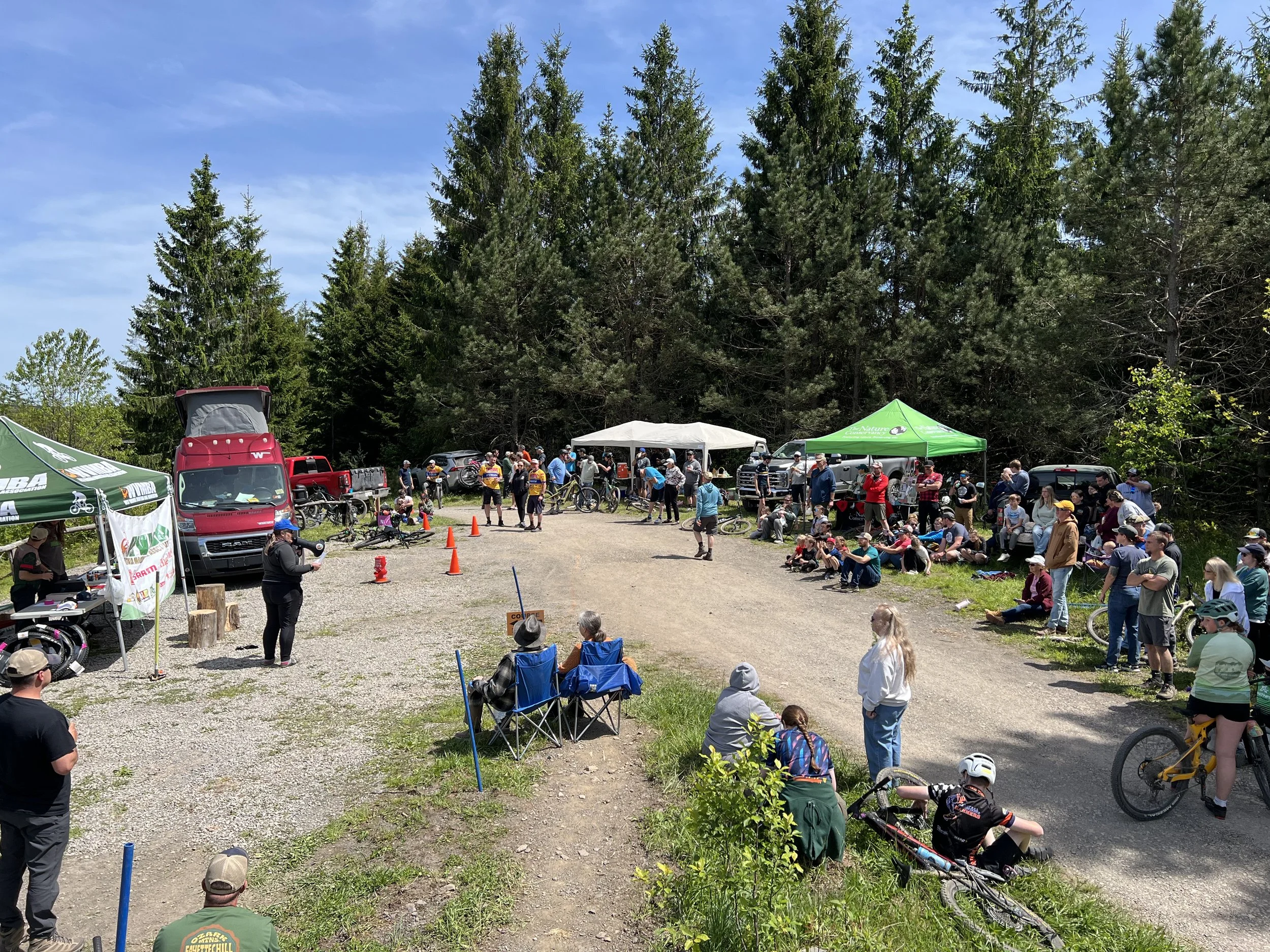 A large outdoor gathering at a mountain bike event with people watching, tents, bikes, and forestry in the background on a sunny day.