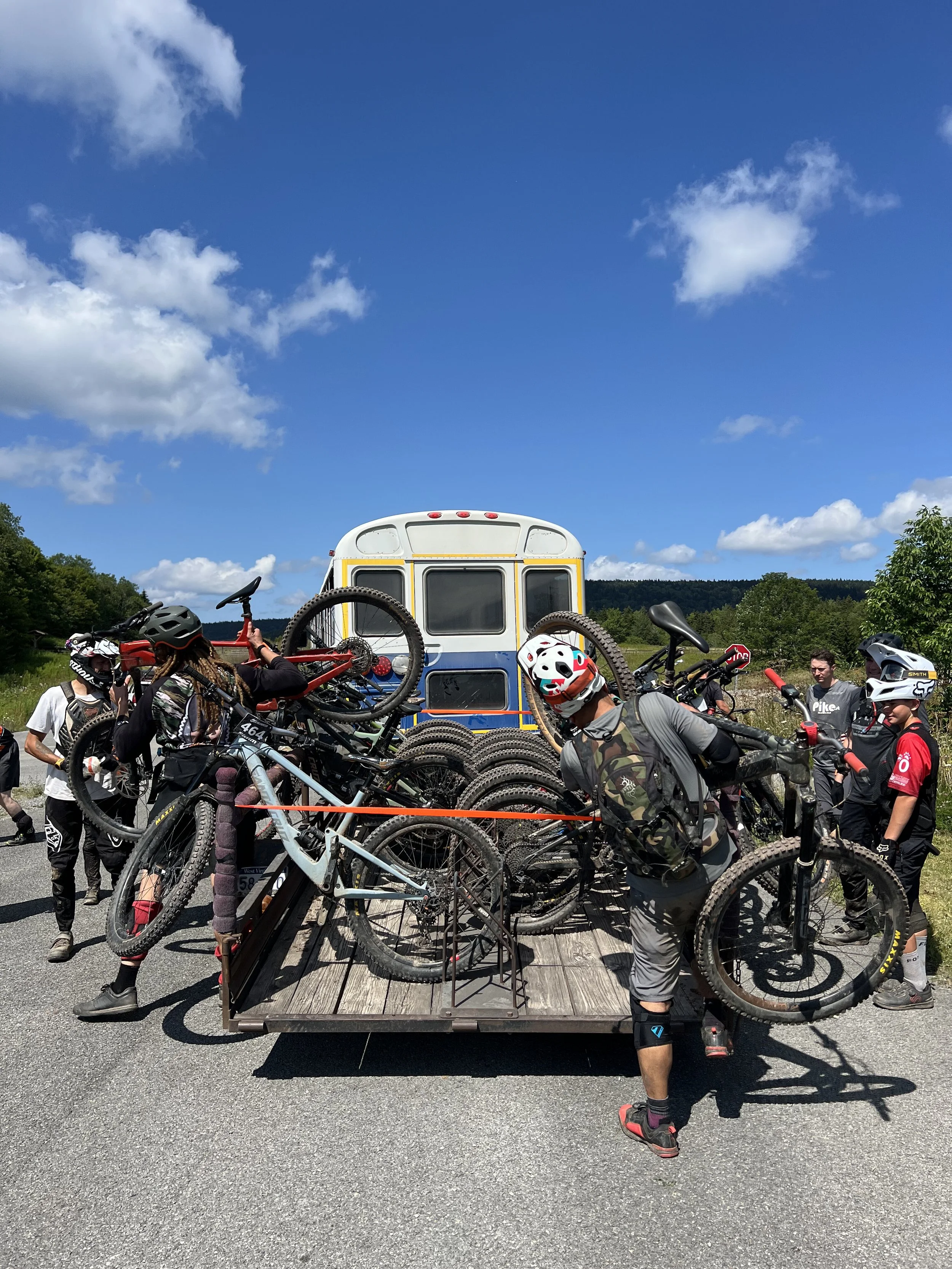 Group of people loading bikes onto a trailer attached to a bus in a scenic outdoor setting.