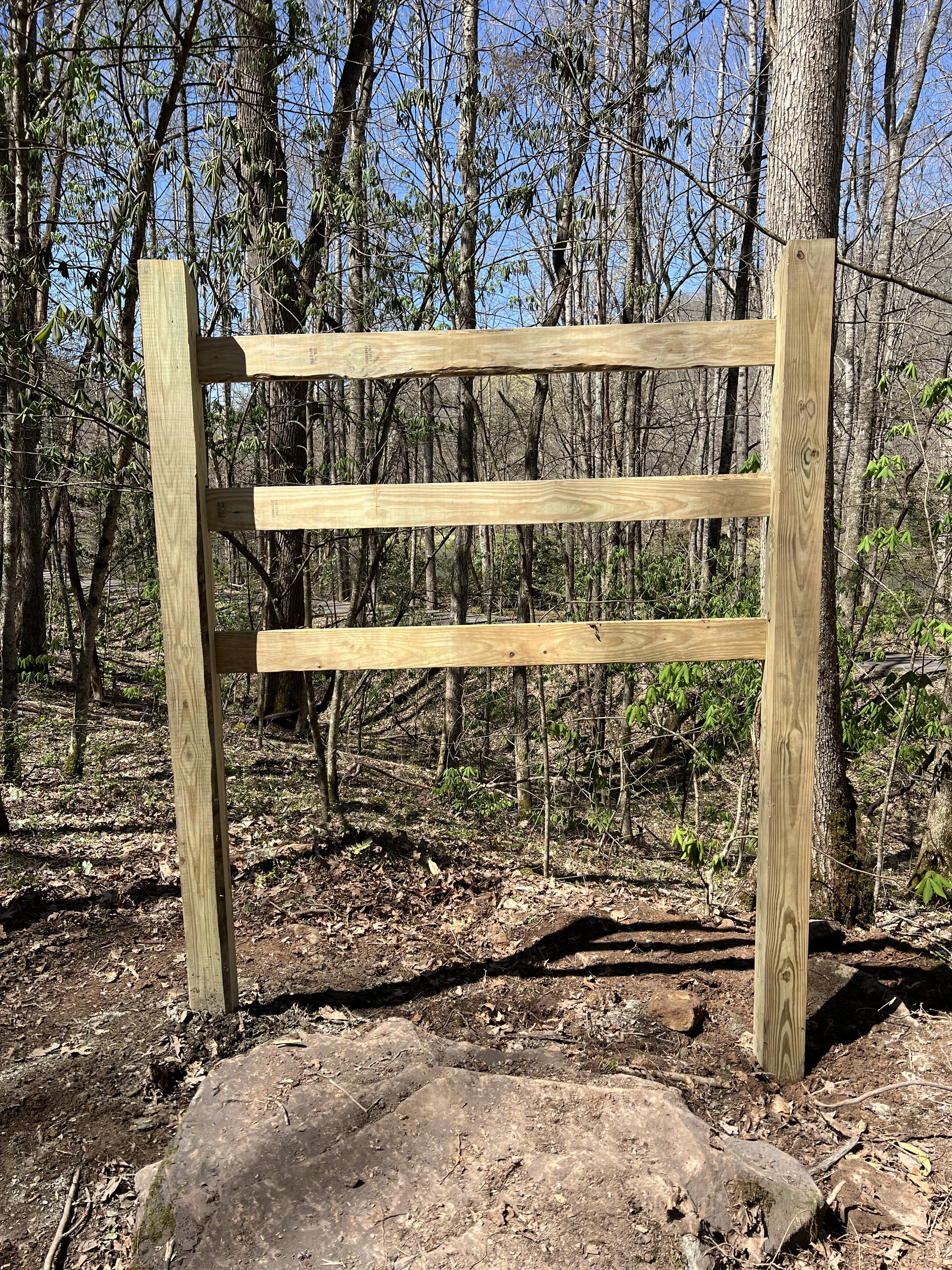 A wooden structure with three horizontal beams supported by vertical posts in a wooded outdoor area with leafless trees and a partly cloudy sky.