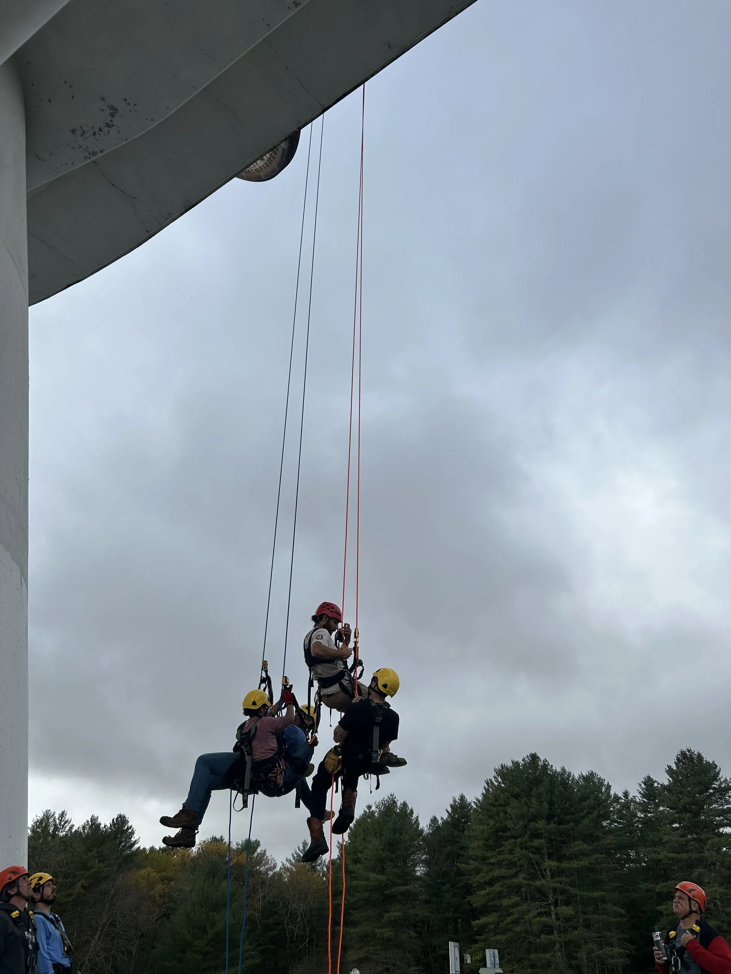 Three people wearing helmets and harnesses are participating in a rescue training exercise, suspended in mid-air by ropes near a large building with a wooded area in the background.