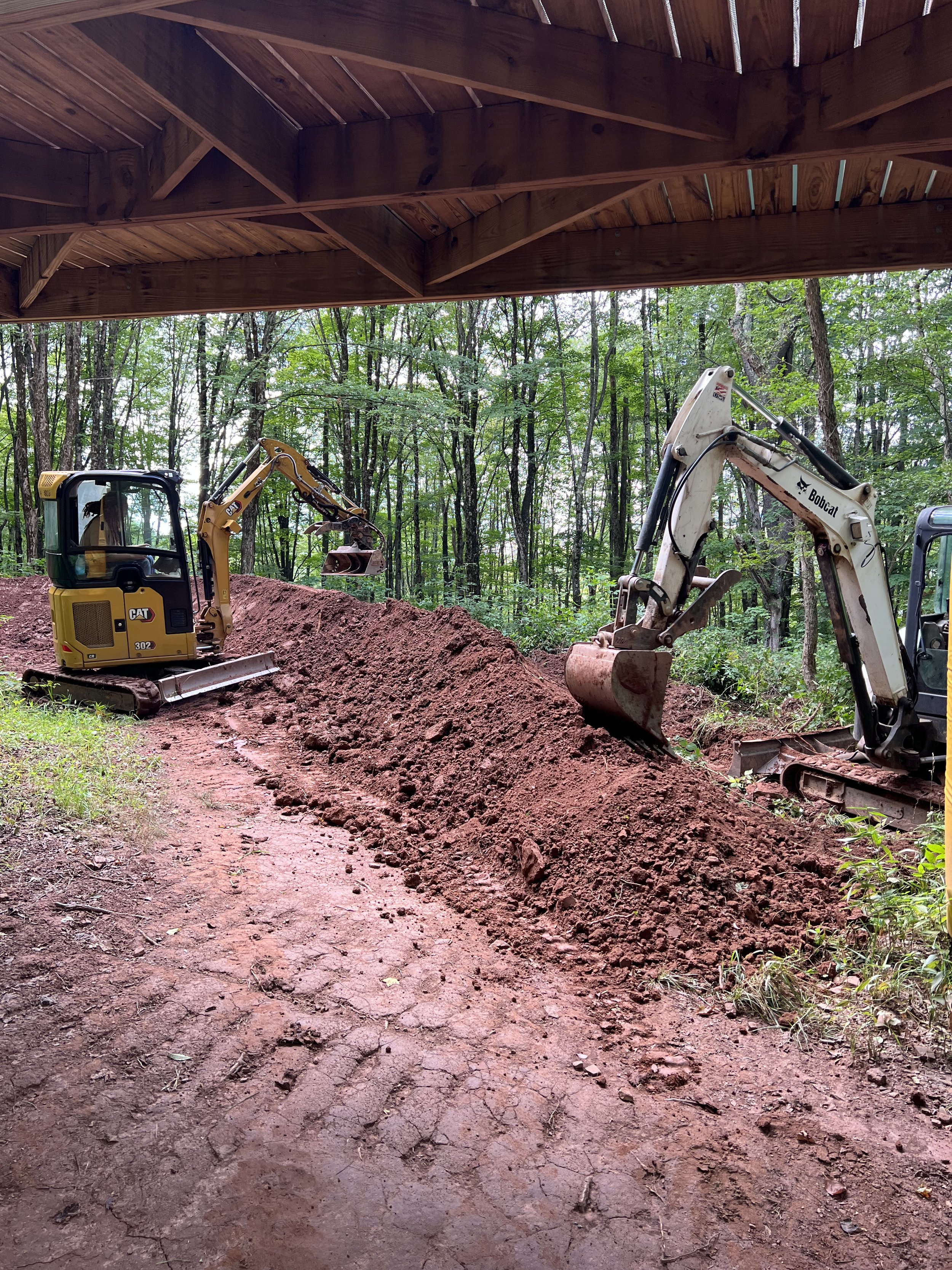 Two excavators on a construction site under a wooden shelter, moving dirt in a wooded area.