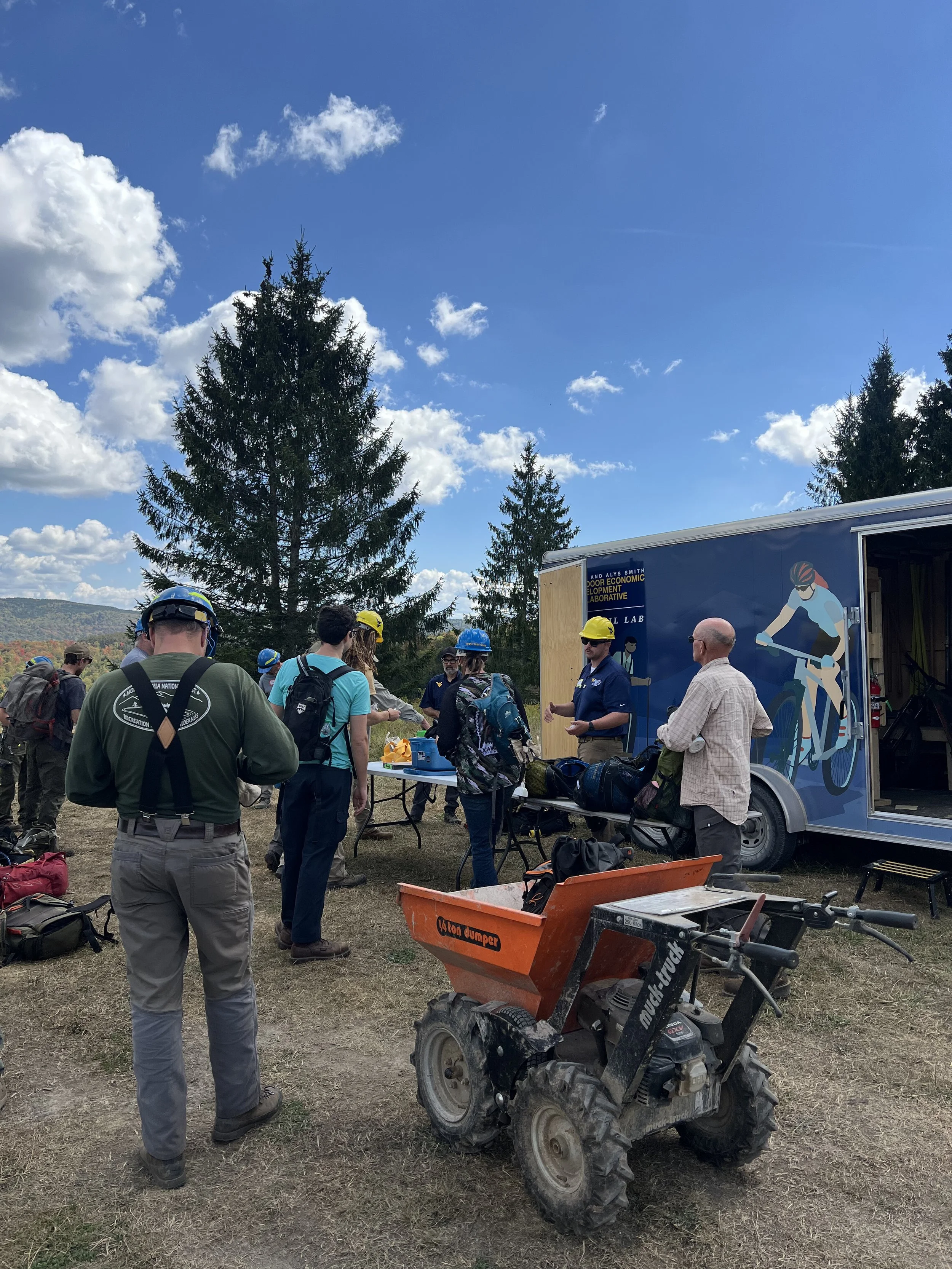 Group of people gathered outdoors near a mobile lab trailer, with trees and blue sky overhead. Some individuals are wearing helmets. There are backpacks, a small orange utility vehicle, and a table with supplies.