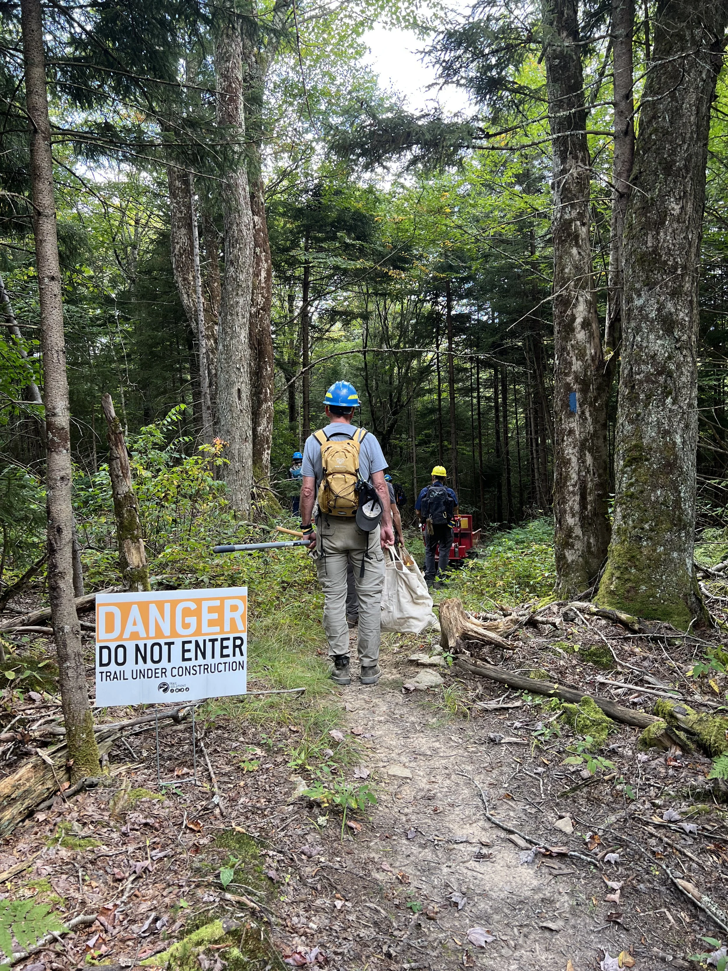 Trail through a forest with a warning sign that says "DANGER DO NOT ENTER TRAIL UNDER CONSTRUCTION"; several workers with helmets and backpacks are walking along the trail.