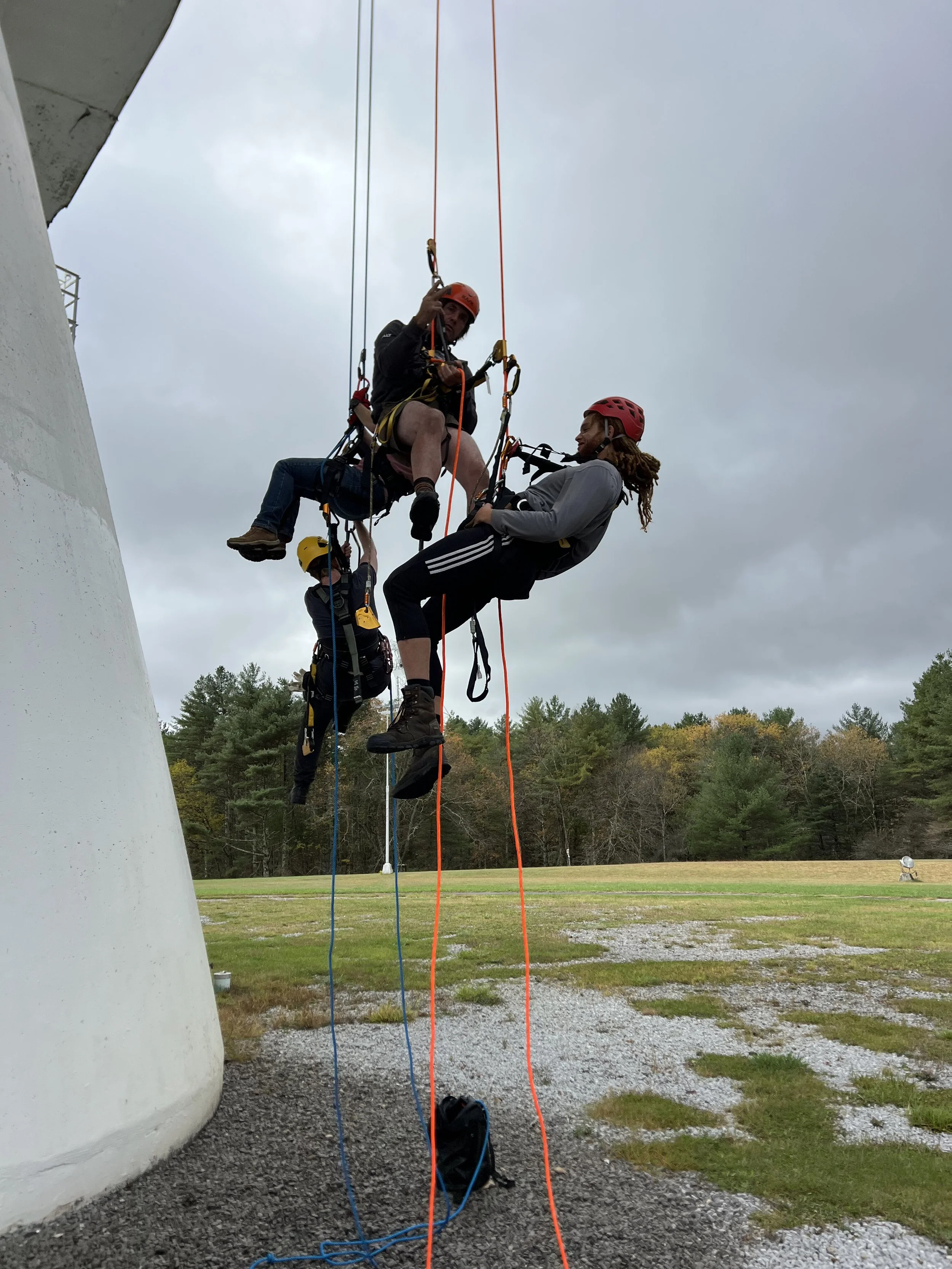 Two individuals in safety harnesses and helmets working at height on climbing equipment near a large white structure, with a backdrop of trees and an overcast sky.