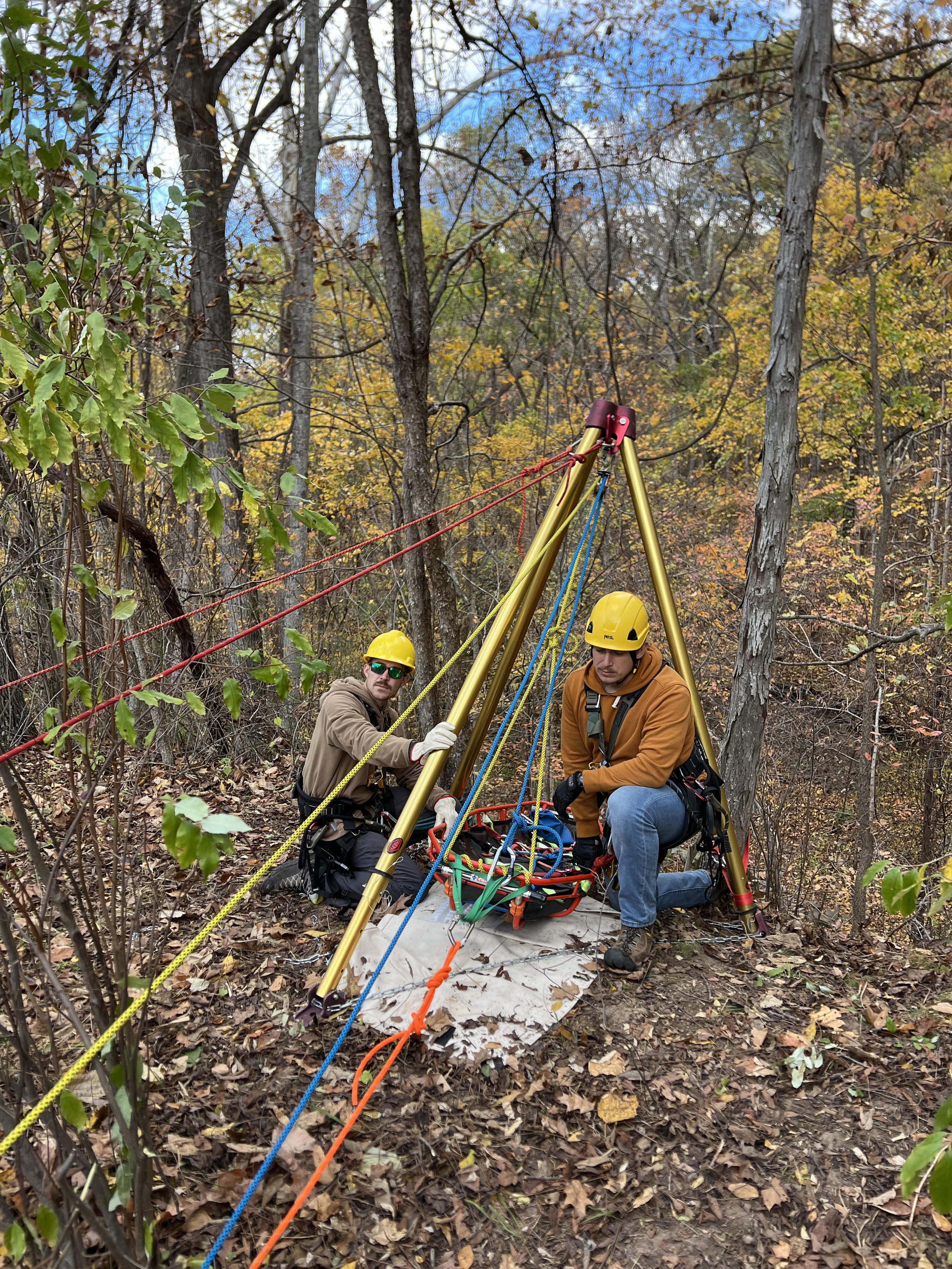 Two men wearing yellow helmets and outdoor gear setting up a construction or scientific apparatus in a forest with autumn leaves on the ground and trees.
